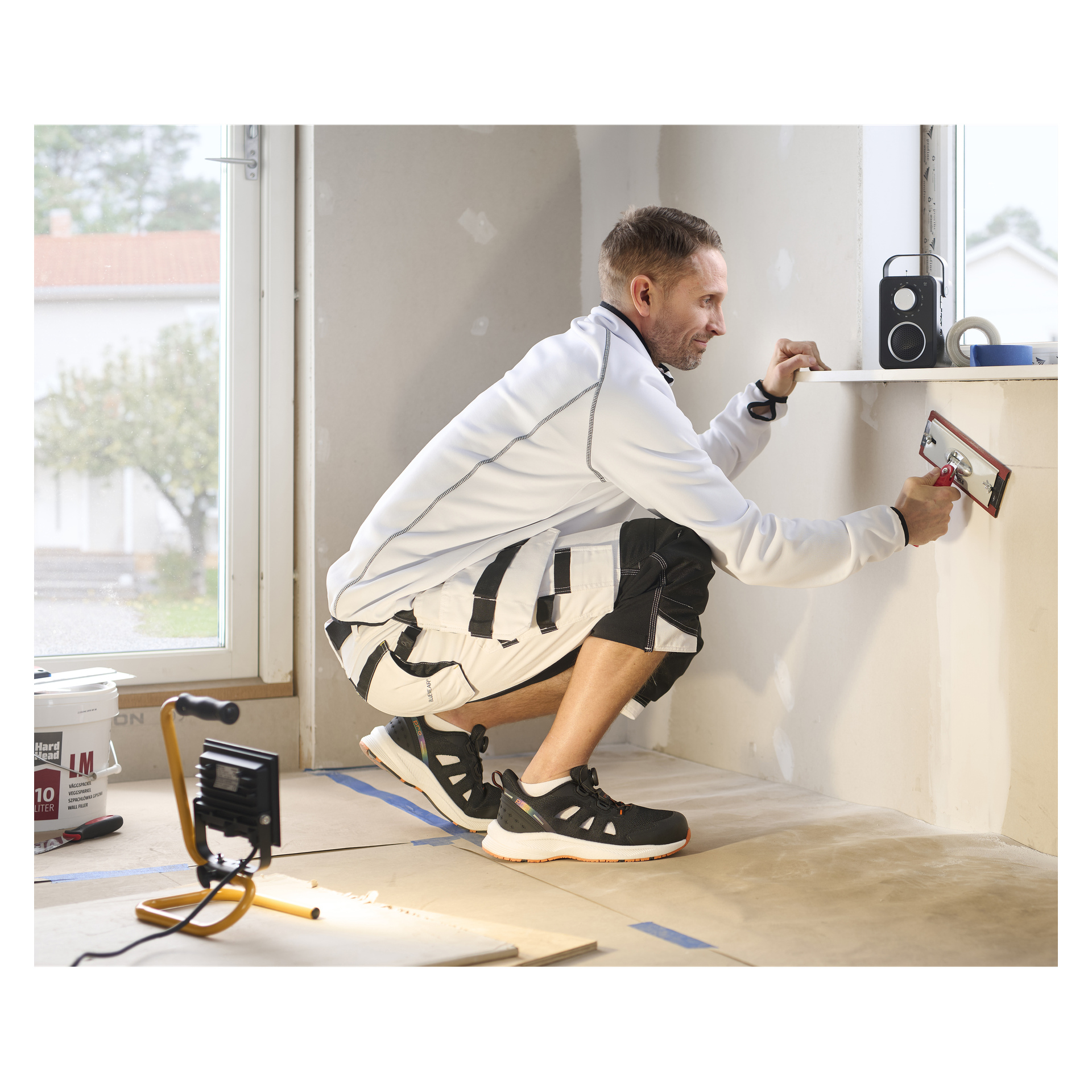 En man i Bluewear Pro Workwear Safety sandaler använder en murslev för att jämna ut puts på en fönsterbräda; Verktyg och lampor finns i närheten.
