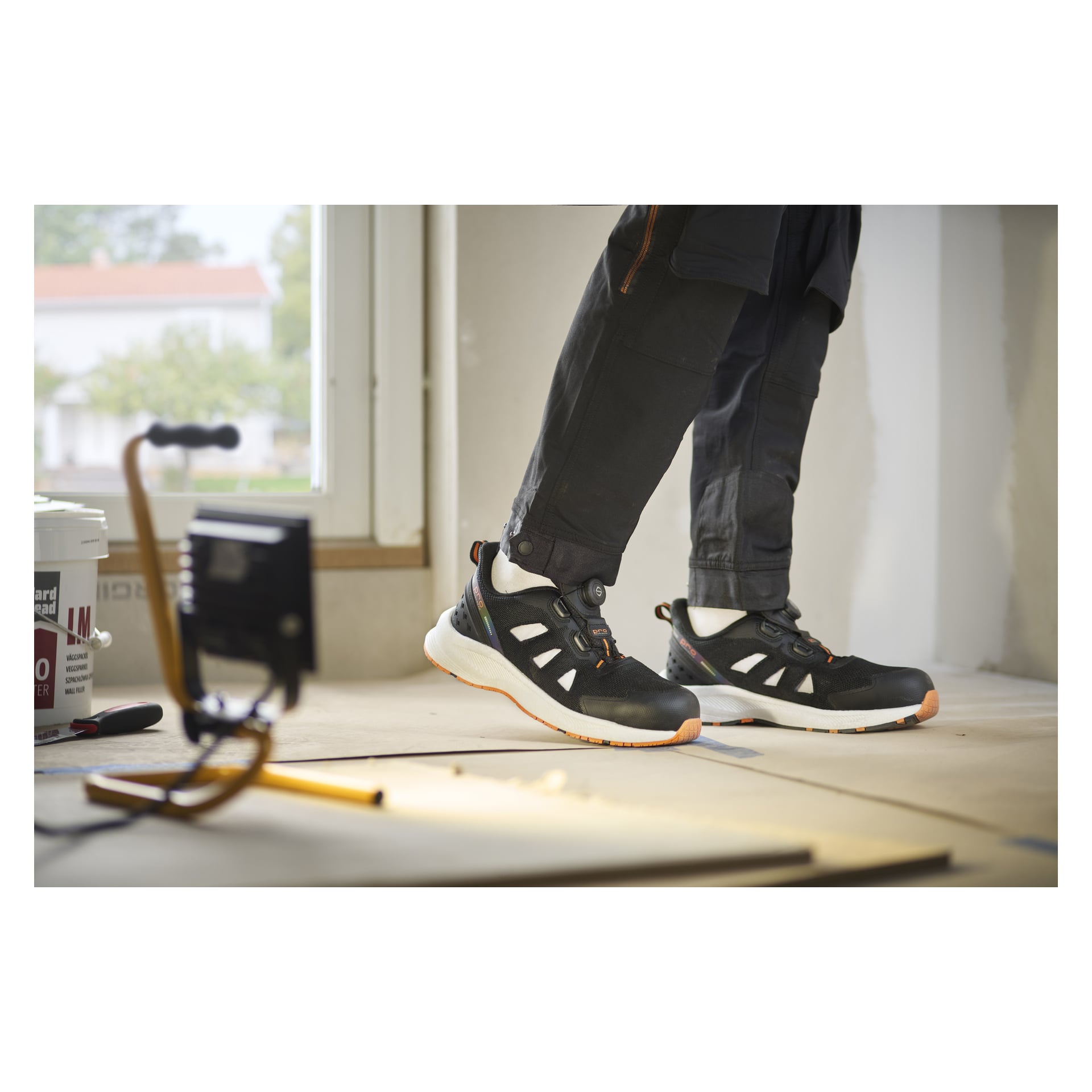 Någon i Bluewear Pro Workwear Austin säkerhetssandaler med snabbsnörning står på kartonggolvet i ett rum som renoveras.