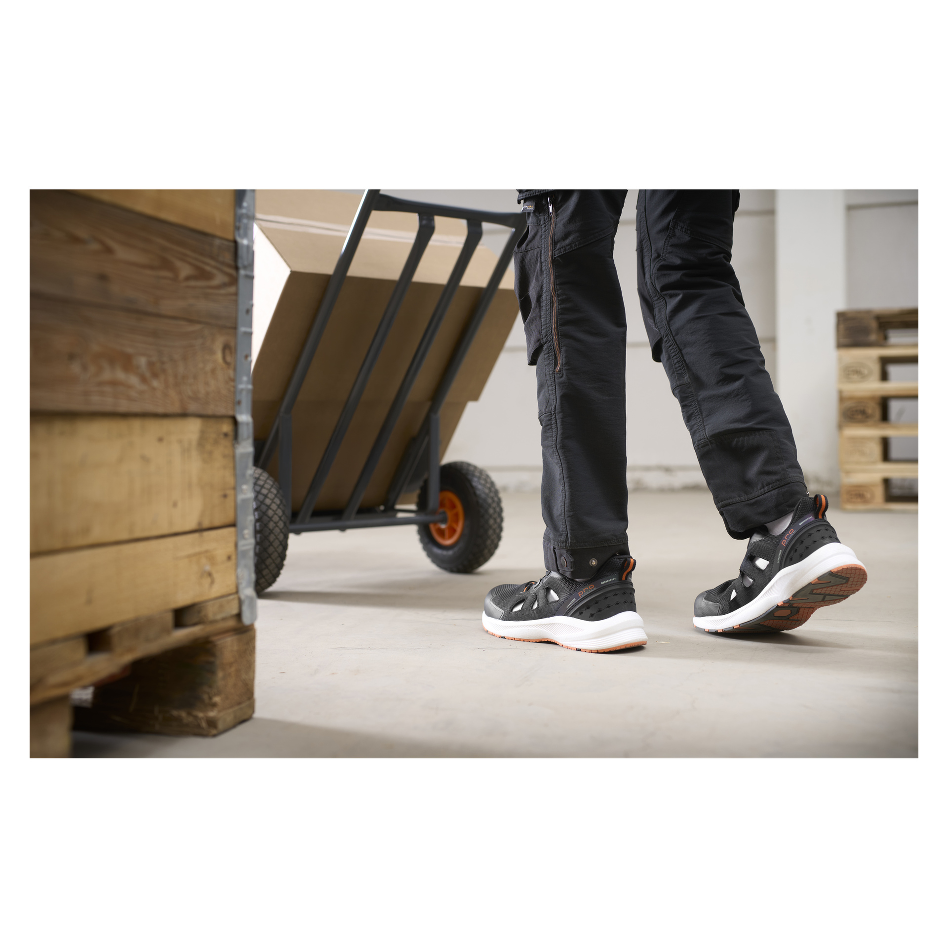 Någon som bär Bluewear Pro Workwear säkerhetssandaler med snabbsnörning från Austin flyttar en stor kartong på en handvagn i ett lager.