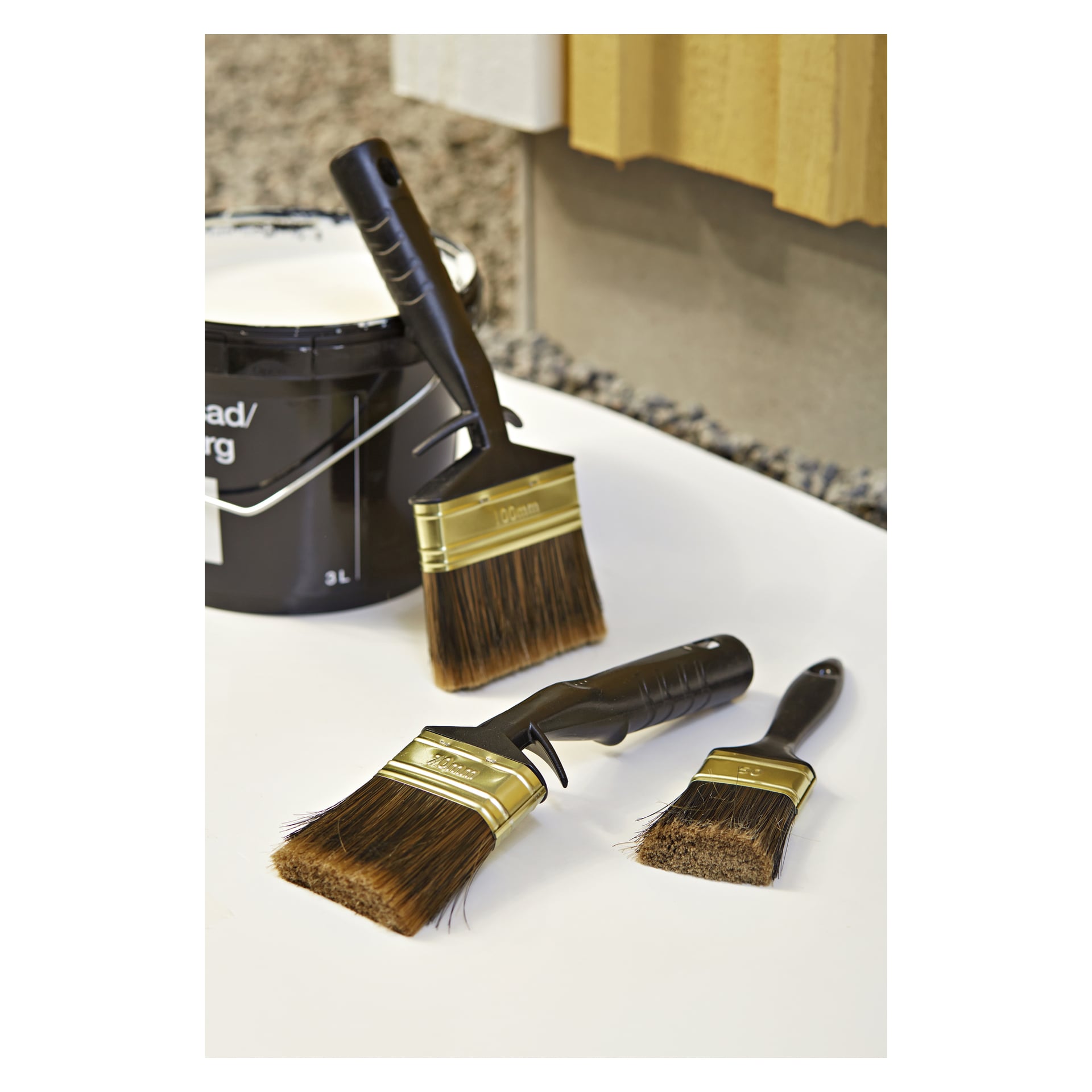 Tre penslar med "Hard Head Brush set 3 pieces" och en svart färgburk visas upp, med en gul vägg och grus i bakgrunden.