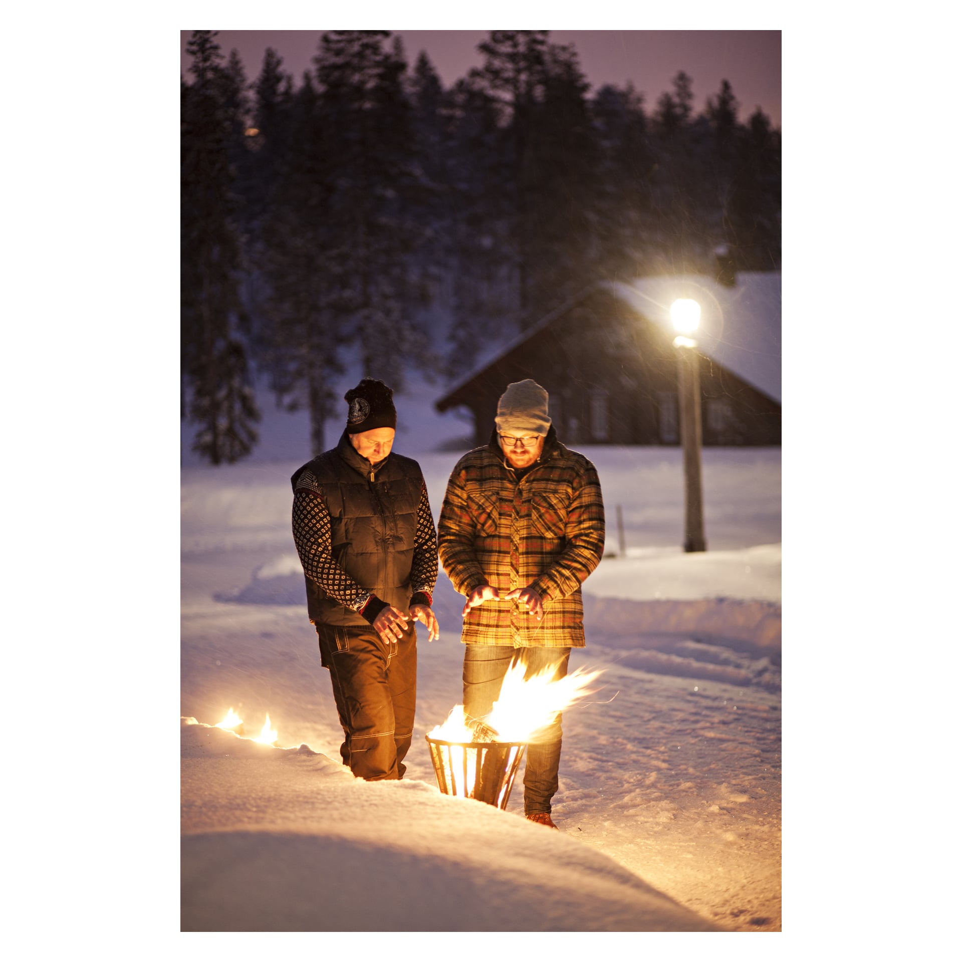 Hämärässä kaksi ihmistä lämmittää käsiään Burns & Barkles -palokorin äärellä lumisessa ulkona mökin, puiden ja katuvalon lähellä.