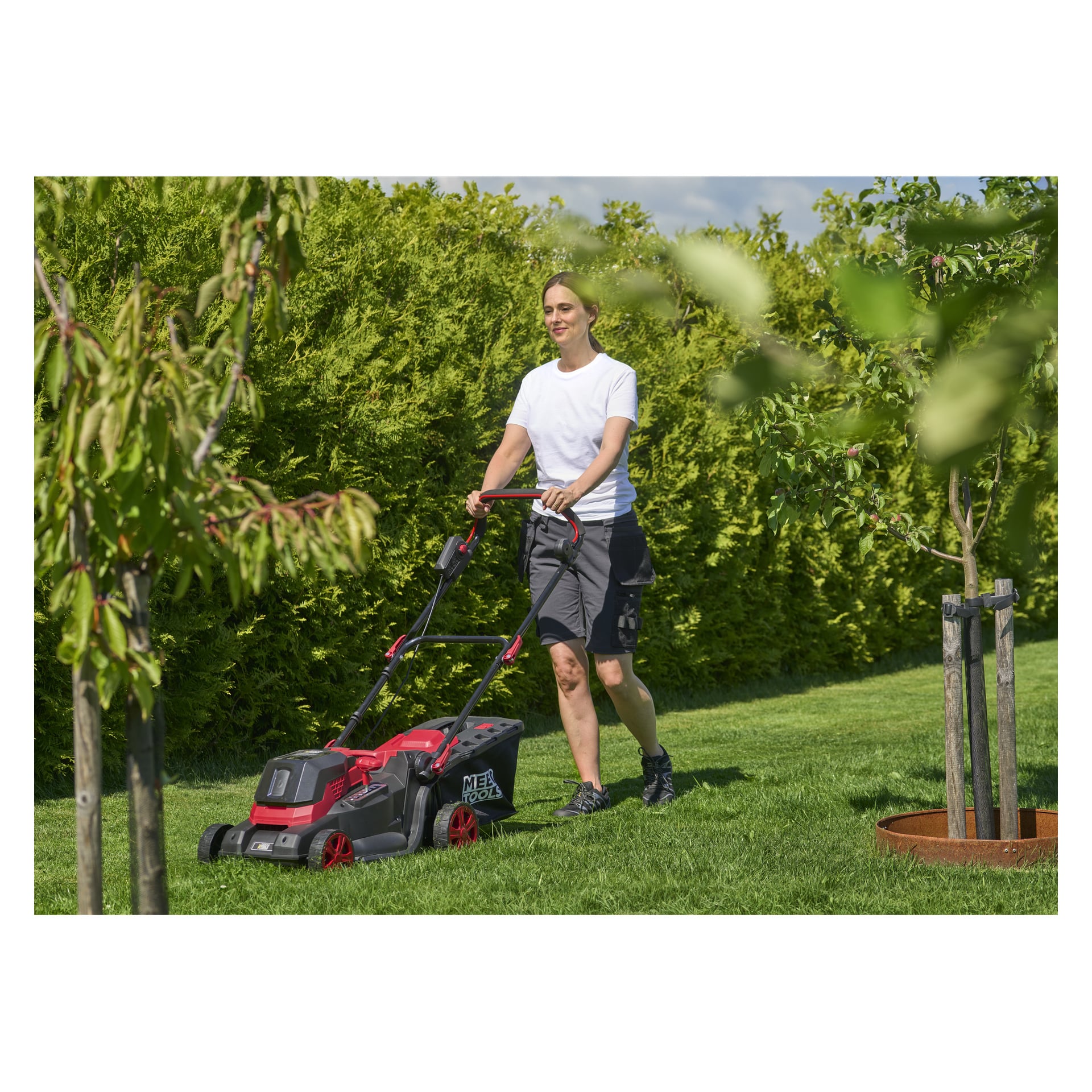 Ktoś w białej koszulce i ciemnych szortach kosi trawnik w ogrodzie Meec Tools LM-18V HOME 300, otoczony drzewami i żywopłotami.