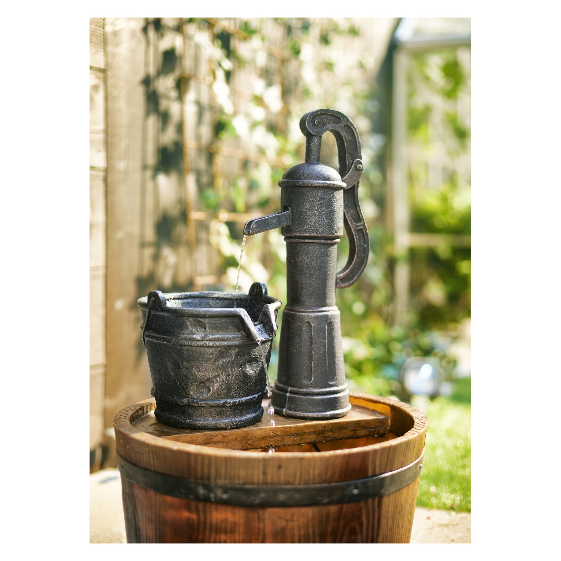 "The Axley Fountain" viser en svart håndpumpe som heller vann i en svart bøtte som hviler på en tønne av tre, omgitt av grønt og sollys.