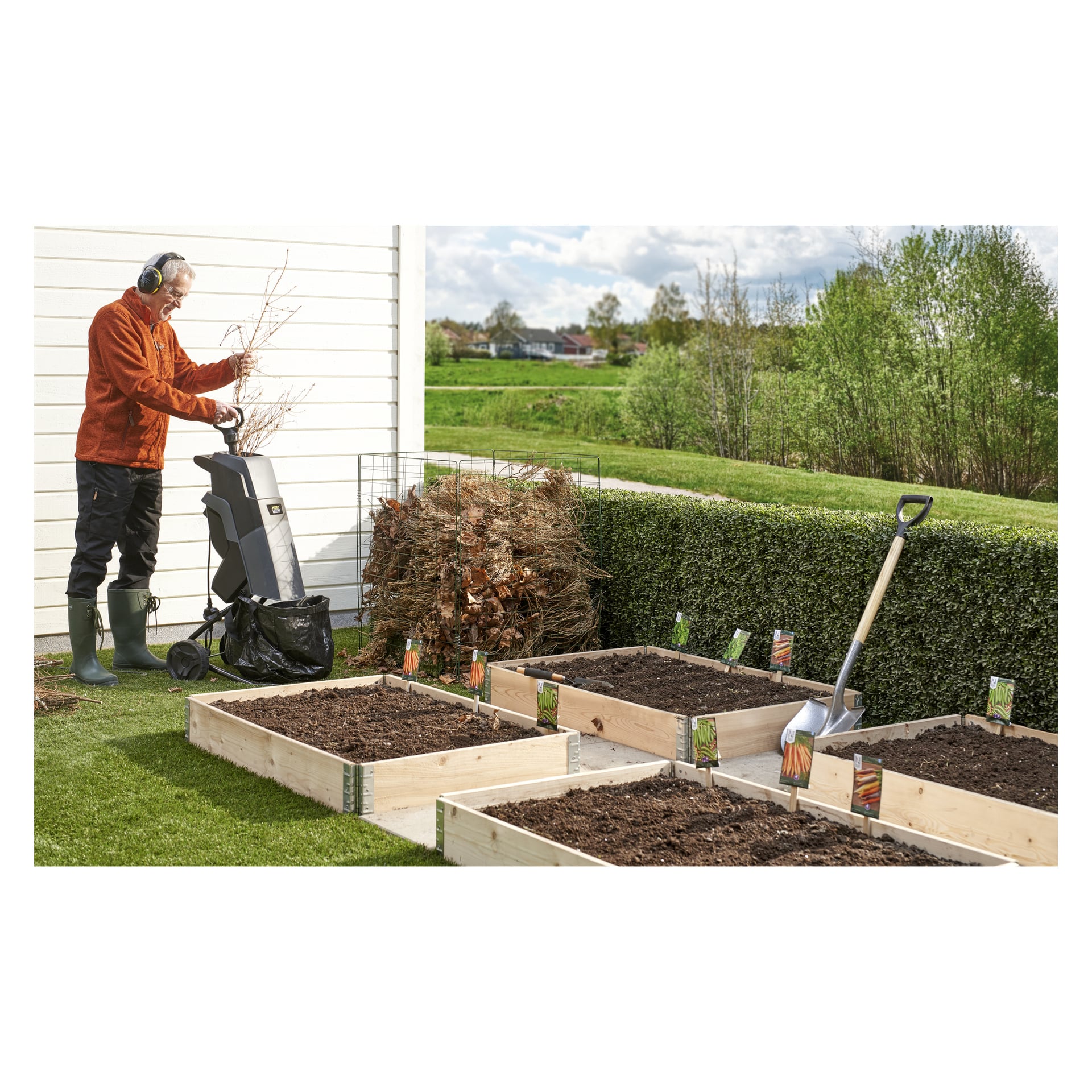 En eldre person i støvler og hodetelefoner mater greiner inn i en Hard Head Compost-makulator. Opphøyde bed og en spade er i nærheten.