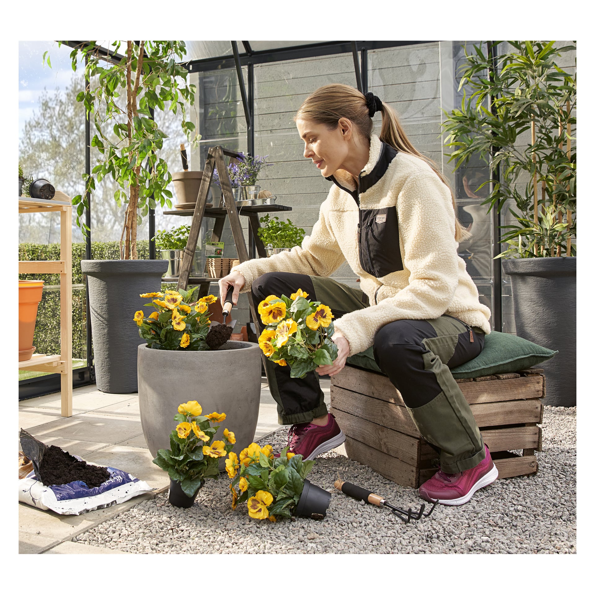 En kvinna planterar gula blommor i en grå kruka med hjälp av Hard Head Planting-setet, sittande på en låda i ett växthus med verktyg och jord i närheten.