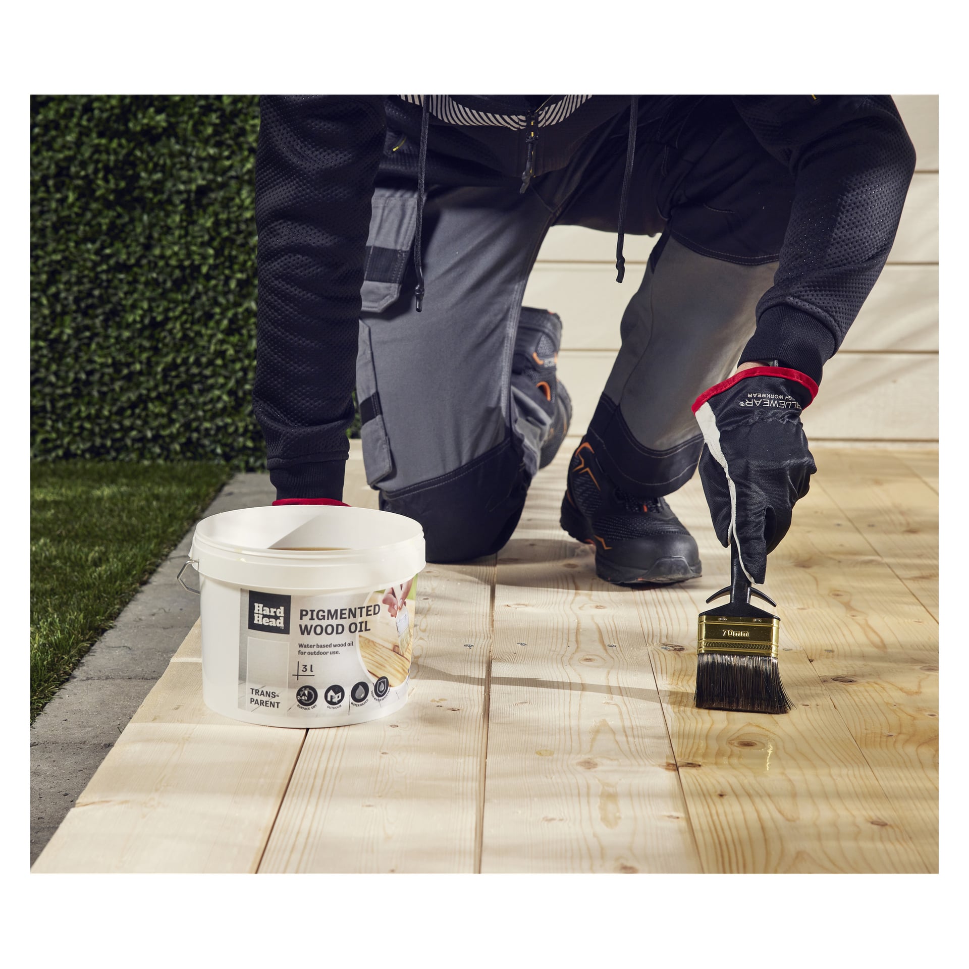 En person i arbetskläder i handske knäböjer på en träaltan och borstar på sig Hard Head Pigmented wood oil från en öppen behållare i närheten.