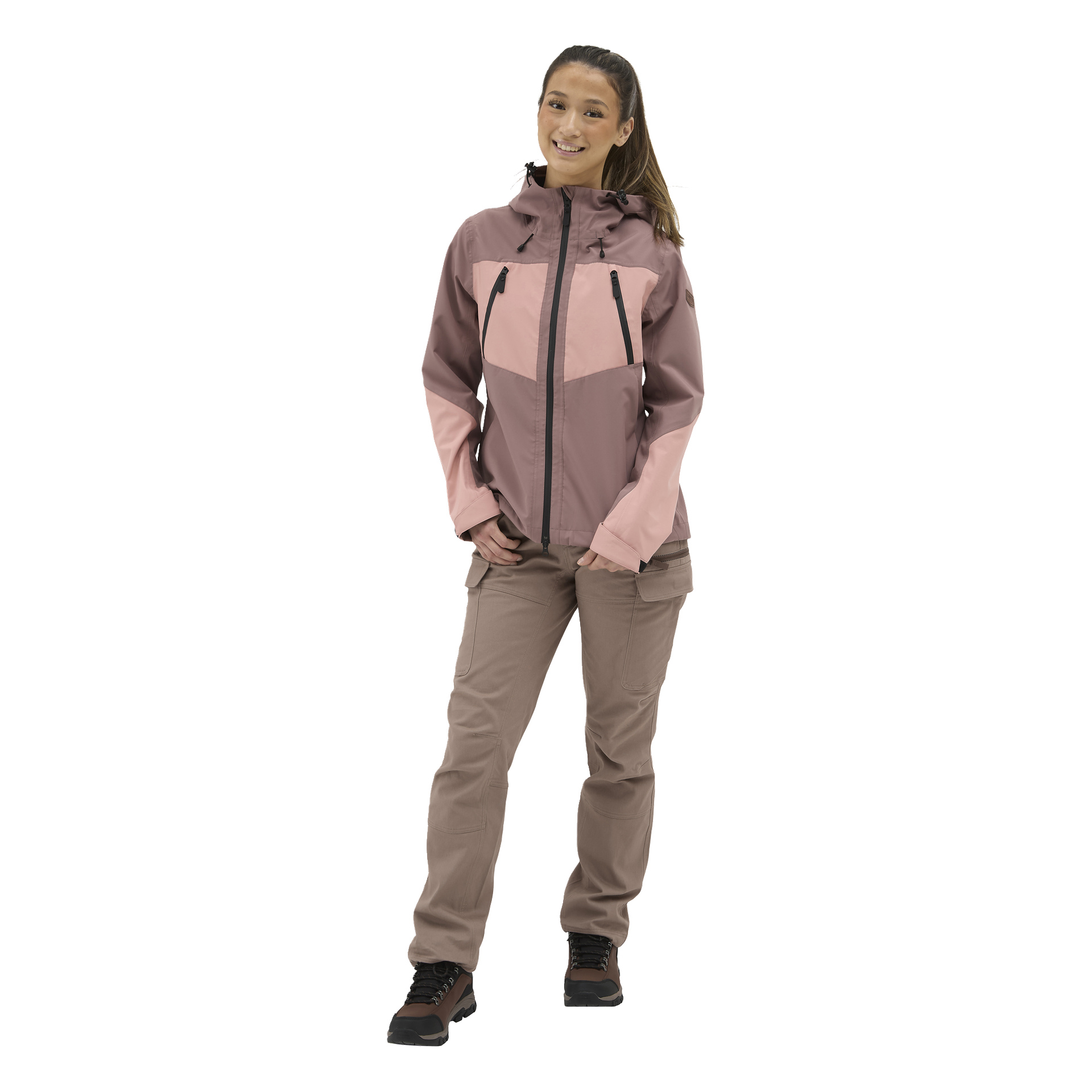 En smilende kvinne med hestehale står vendt fremover, iført lilla og rosa yttertøy, beige bukser og svarte tursko.