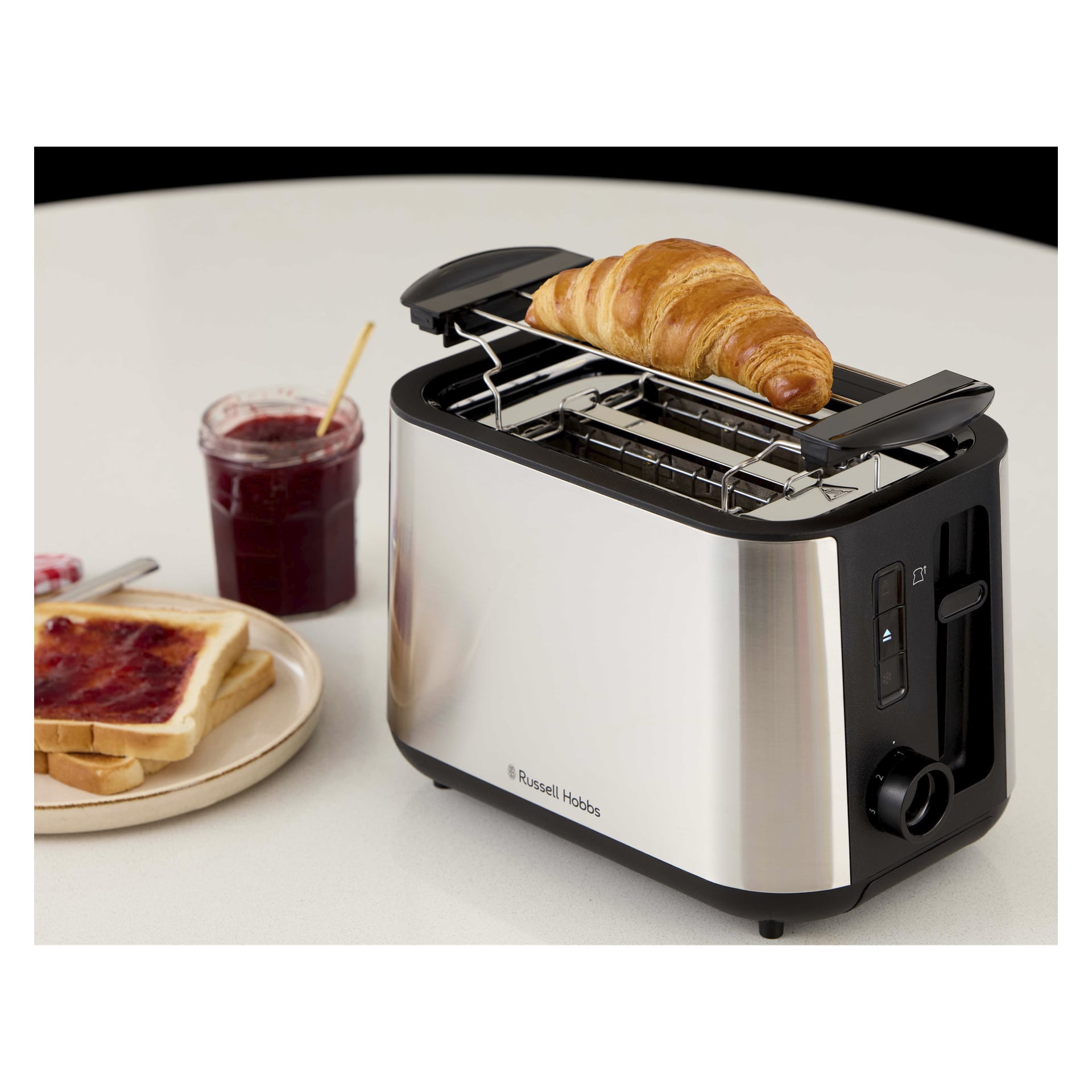 En croissant värms på översta gallret i en Russell Hobbs-brödrost, med sylttoast och en öppen burk sylt med en sked i närheten.
