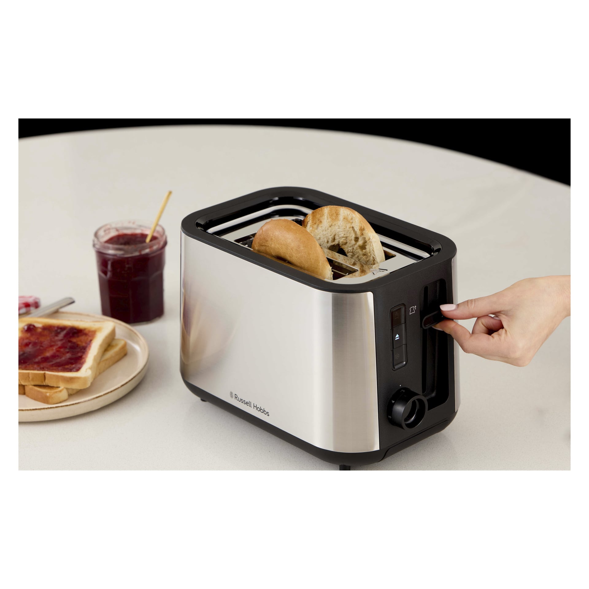 En hand trycker på spaken på en Russell Hobbs tvåskivad brödrost med bröd. Bredvid ligger rostat bröd med sylt, en smörkniv och en syltburk.