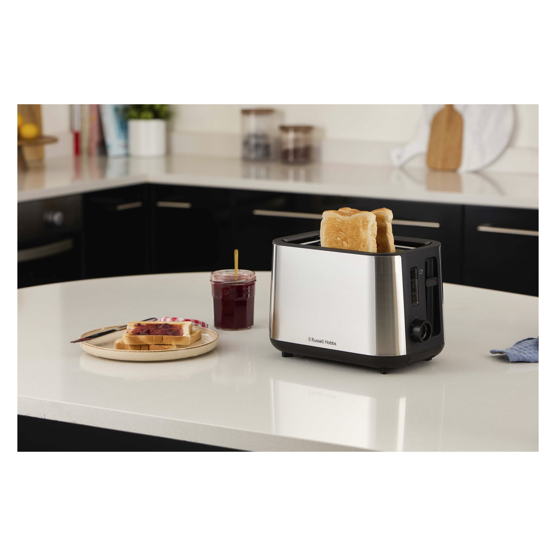 En Russell Hobbs Heaton tvåskivad brödrost på köksbänken med bröd som rostas, bredvid rostat bröd, sylt och andra köksartiklar.