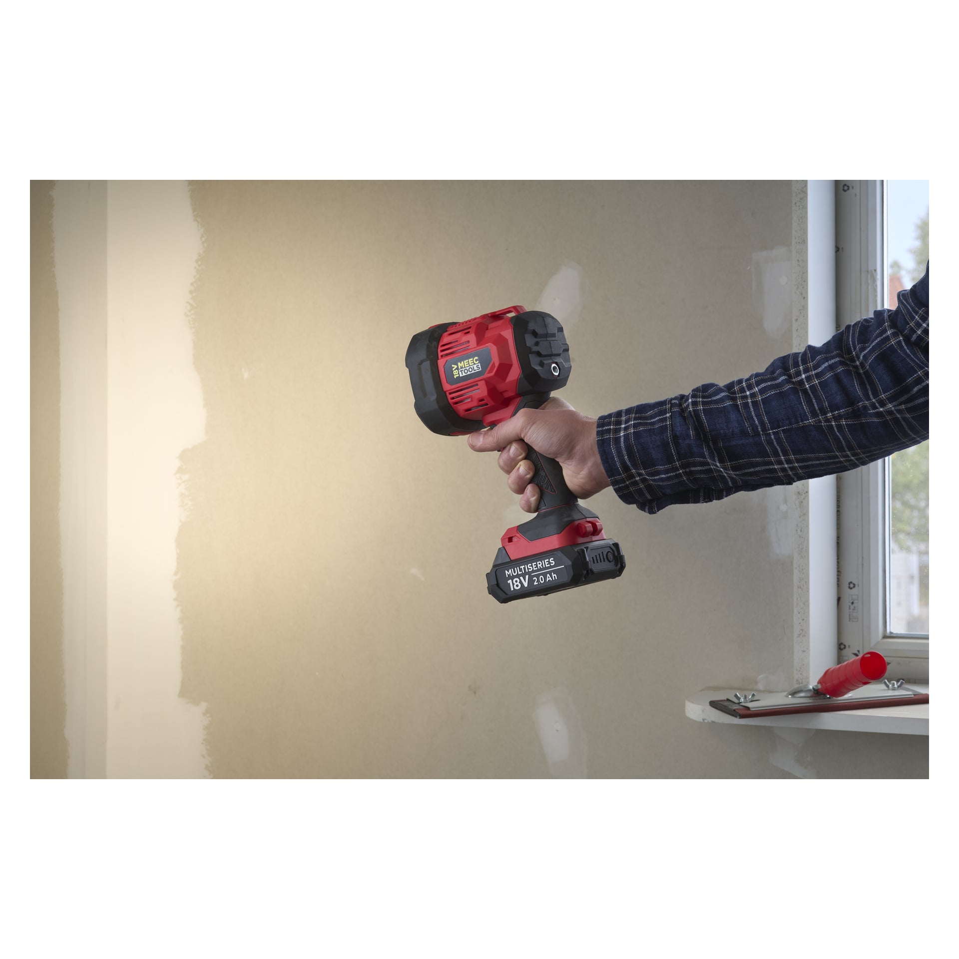 Ktoś w niebieskiej koszuli w kratę trzyma reflektor LED Meec Tools LD-18V HOME 300 przy beżowej ścianie z oknem i narzędziami za nimi.