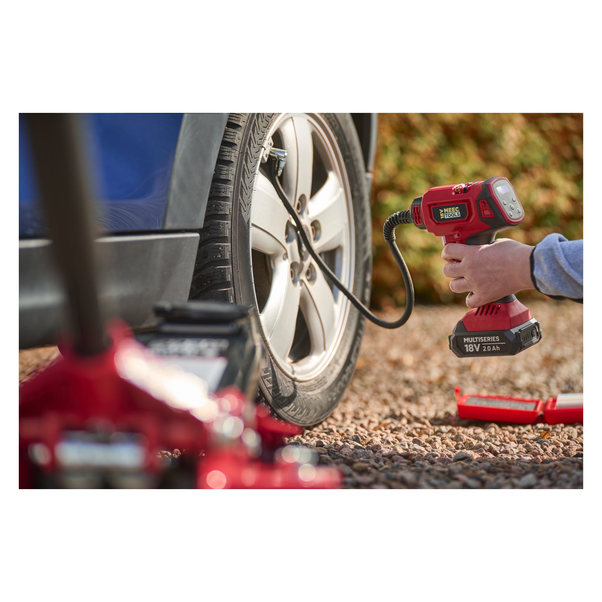 Joku täyttää auton renkaan soralla Meec Tools Multiseries Mini -kompressorilla; Tunkki ja työkalupakki ovat edessä.