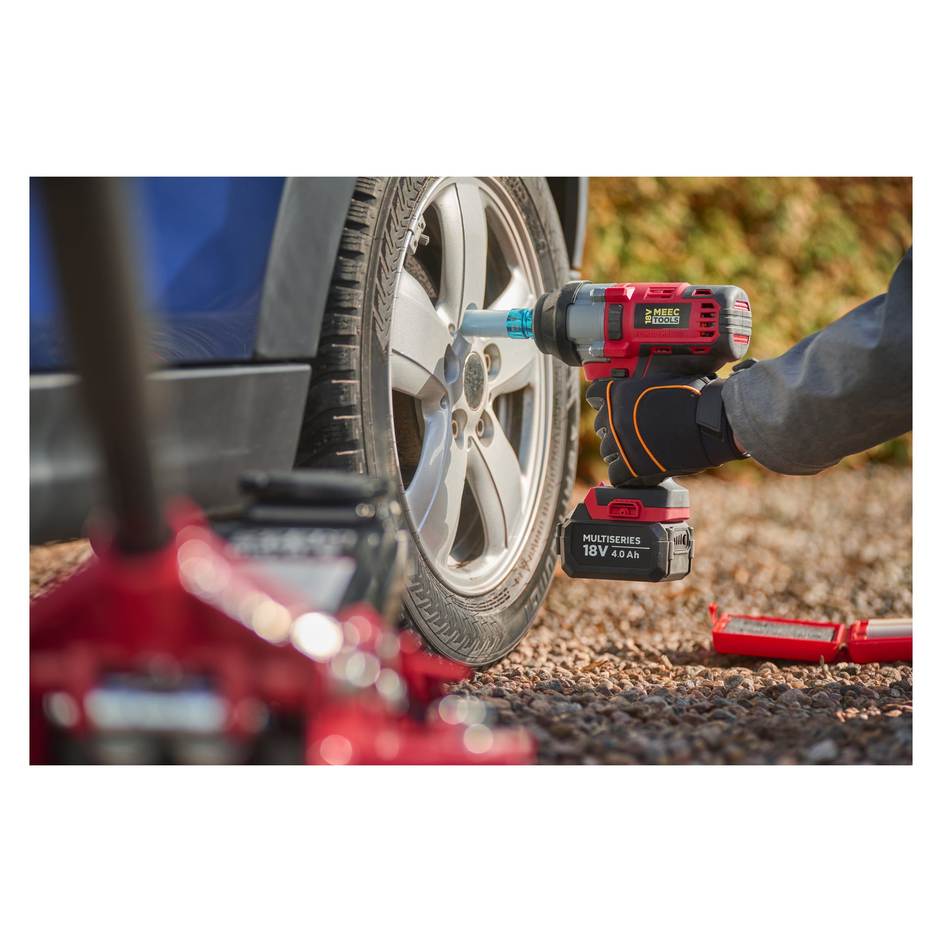 Ktoś obsługuje czerwono-klucz udarowy Meec Tools IW-18V WORK 700 na nakrętkach kół samochodowych, narzędziach rozrzuconych w pobliżu na żwirze.