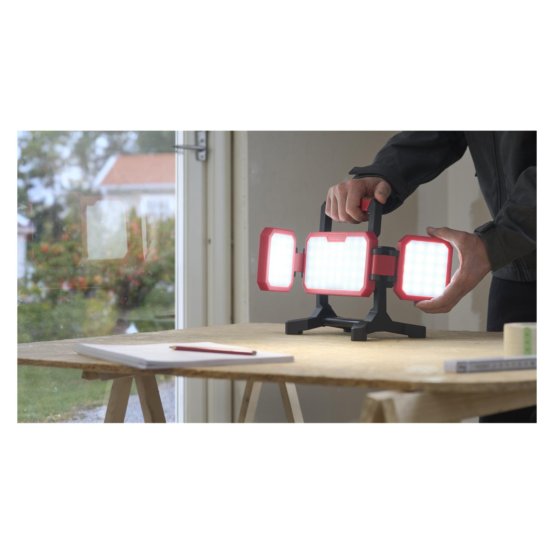 Osoba ustawia lampę Meec Tools Multiseries LD-18V WORK 700 na drewnianym stole przy oknie z notatnikiem, długopisem i taśmą.