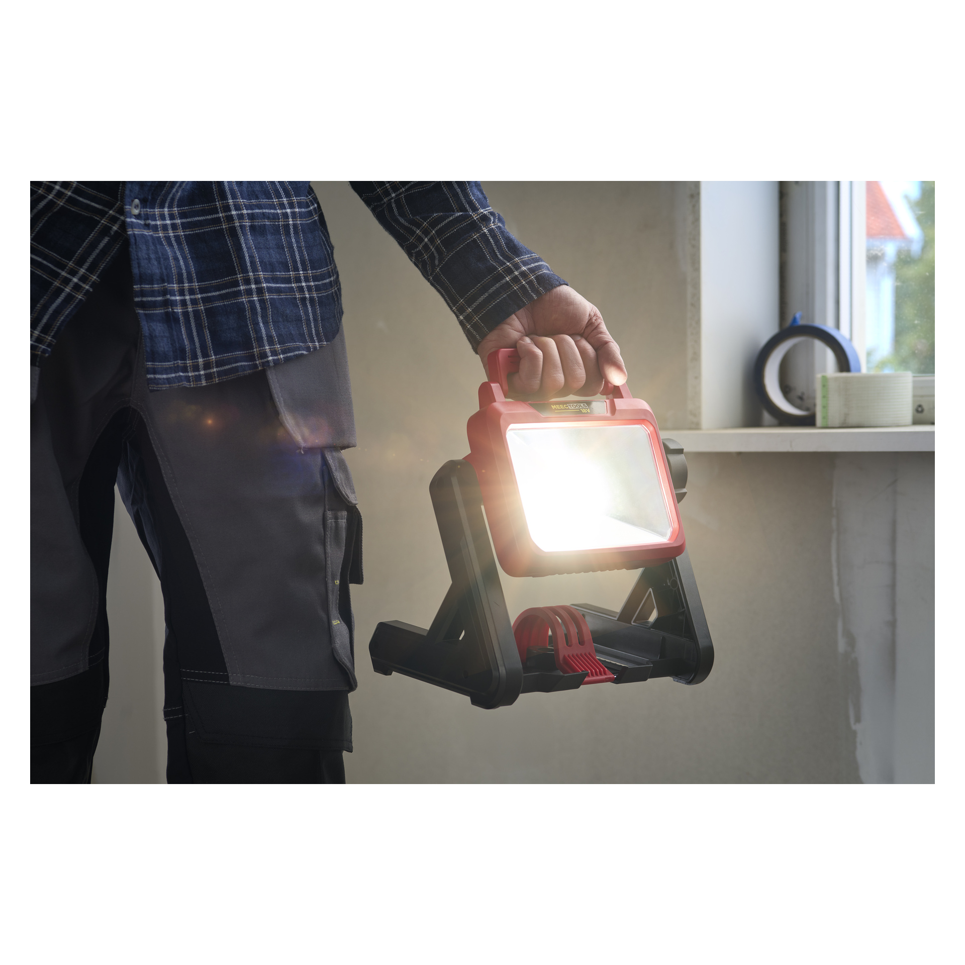En person i grå arbetsbyxor och rutig skjorta håller i en batterilampa från Meec Tools med rött handtag, stående vid en fönsterbräda.