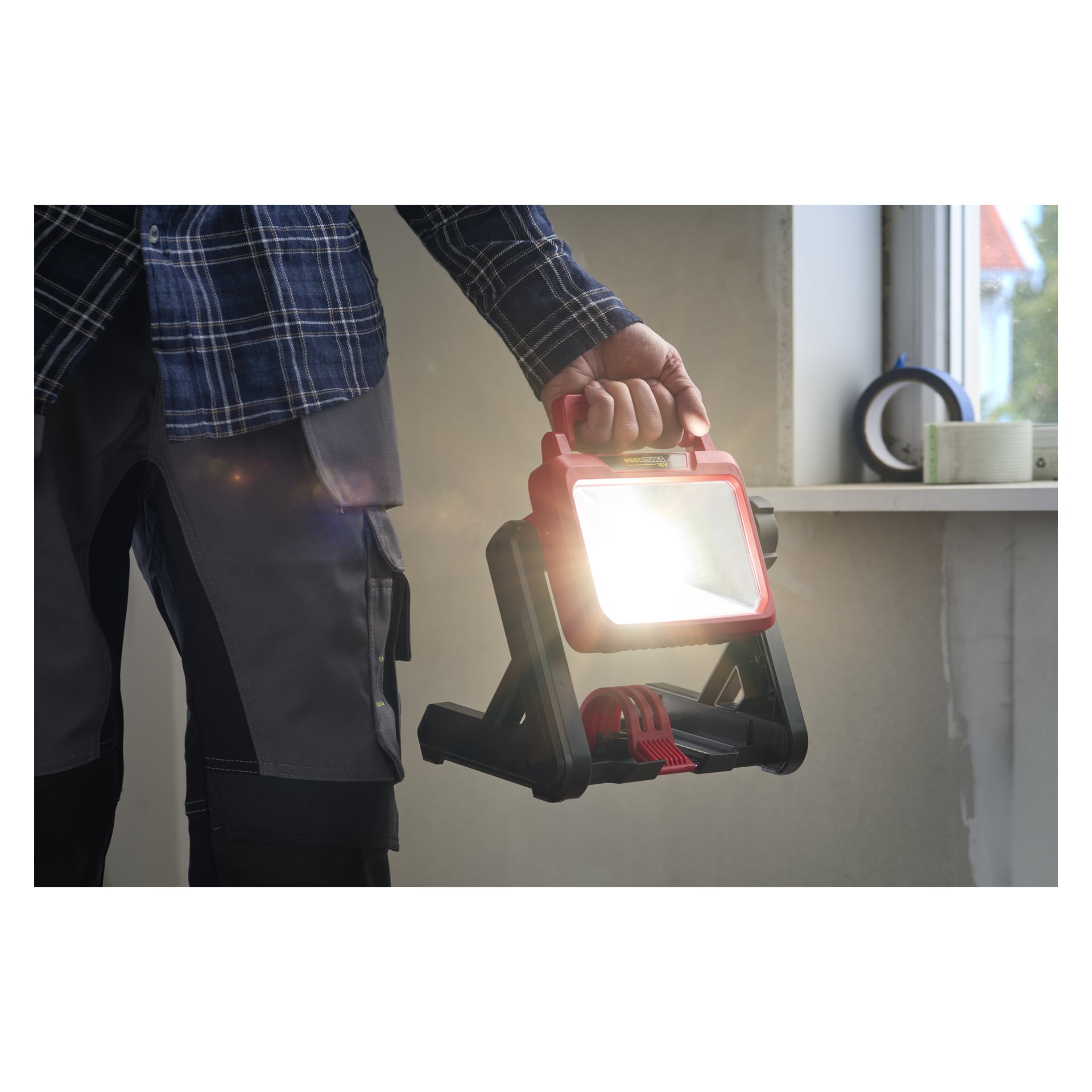 En person i grå arbetsbyxor och rutig skjorta håller i en batterilampa från Meec Tools med rött handtag, stående vid en fönsterbräda.