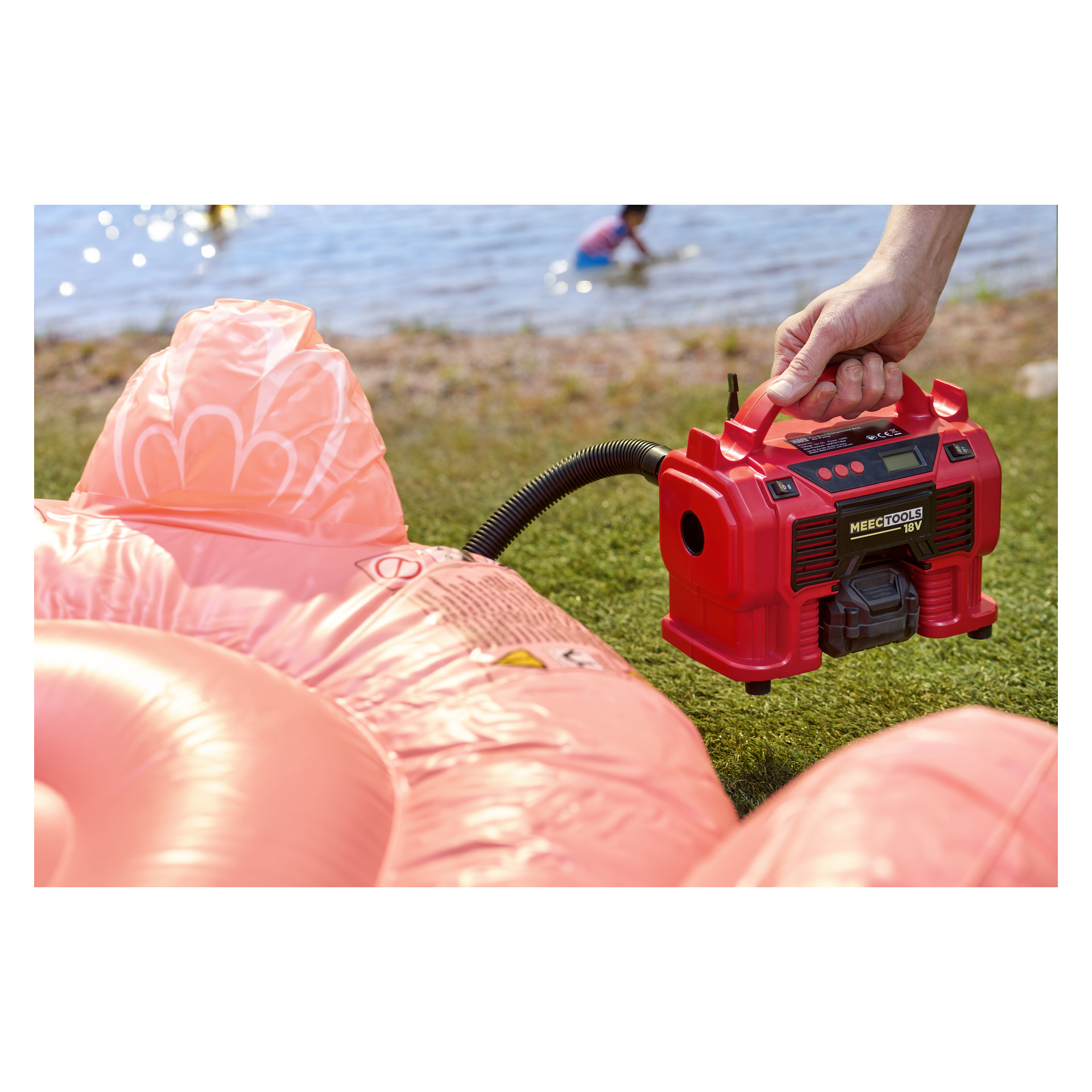 Jemand bläst einen großen rosa Schwimmschwimmer mit einer roten elektrischen Pumpe auf das Gras am See, während Kinder im Wasser dahinter spielen.