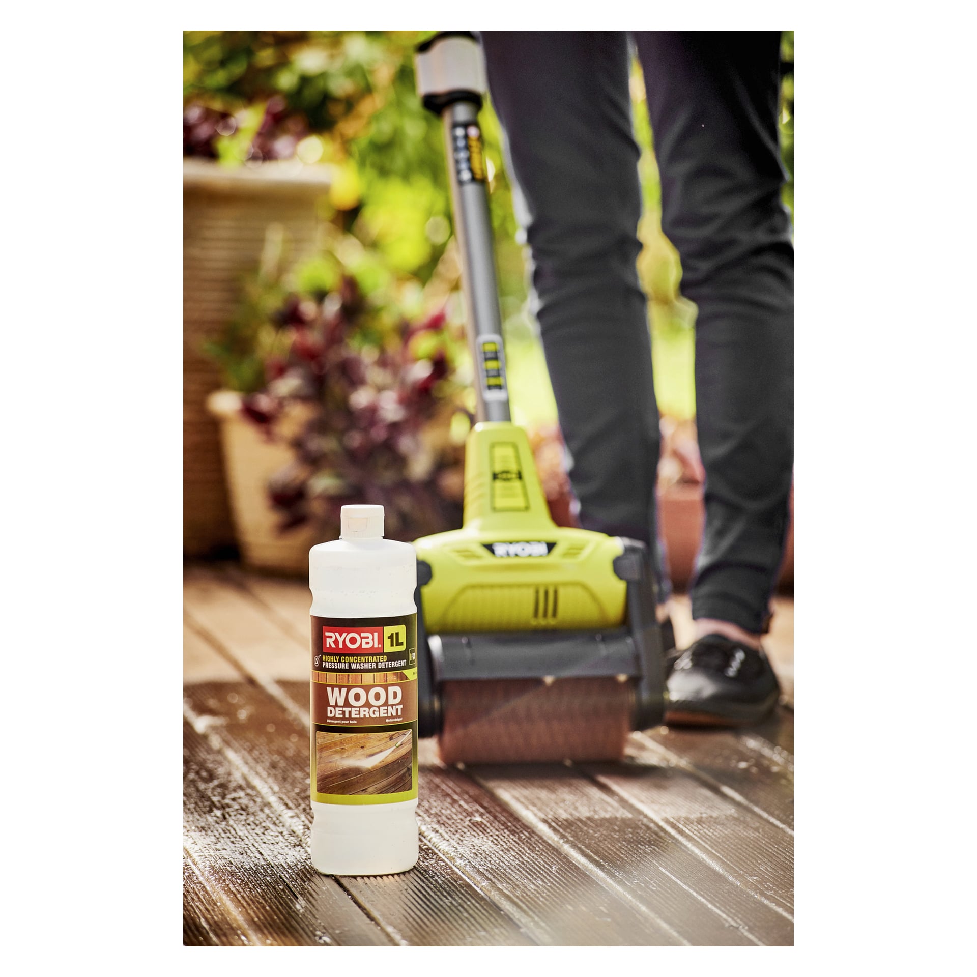 En Ryobi One+ uteplatsrengörare RY18PCB-140W rengör en träaltan, med en flaska Ryobi 1L trätvättmedel och krukväxter i närheten.