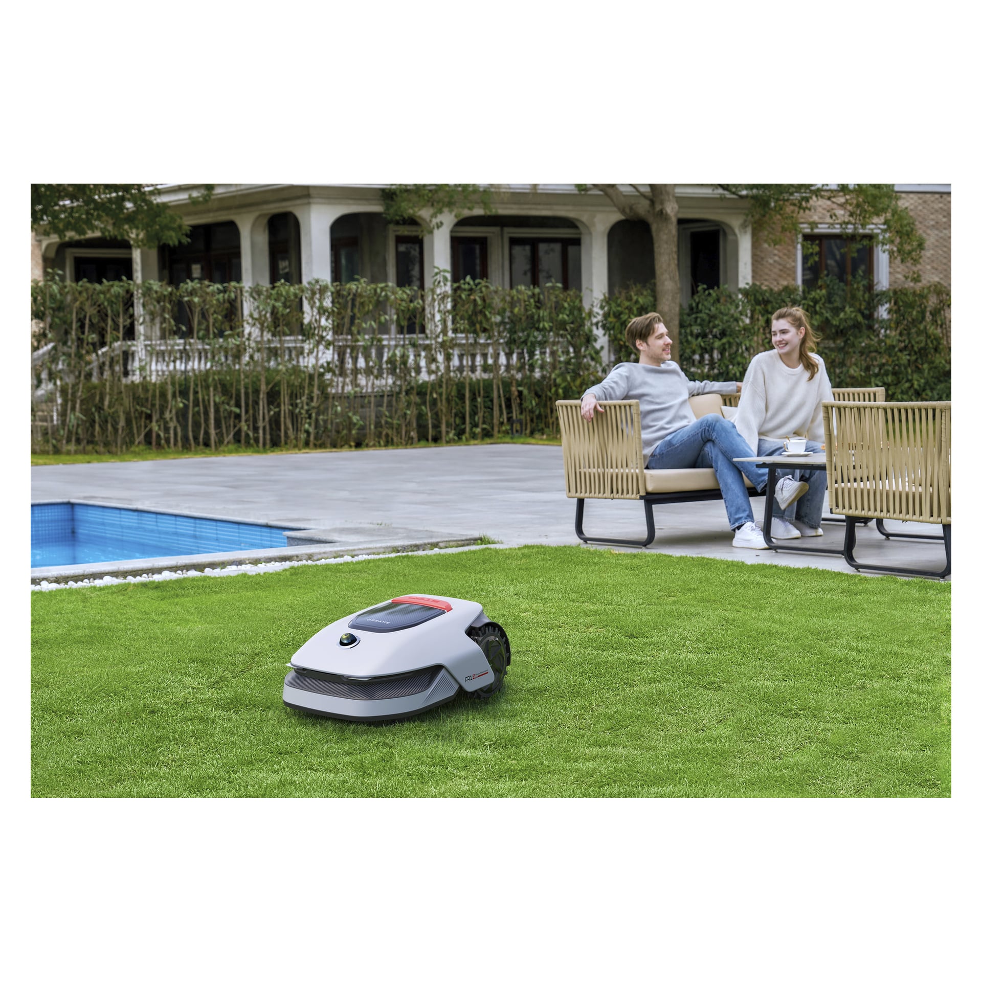 En Dreame-robotgräsklippare klipper gräset vid en pool medan två personer slappar på utemöbler, med ett stort hus och buskar bakom.