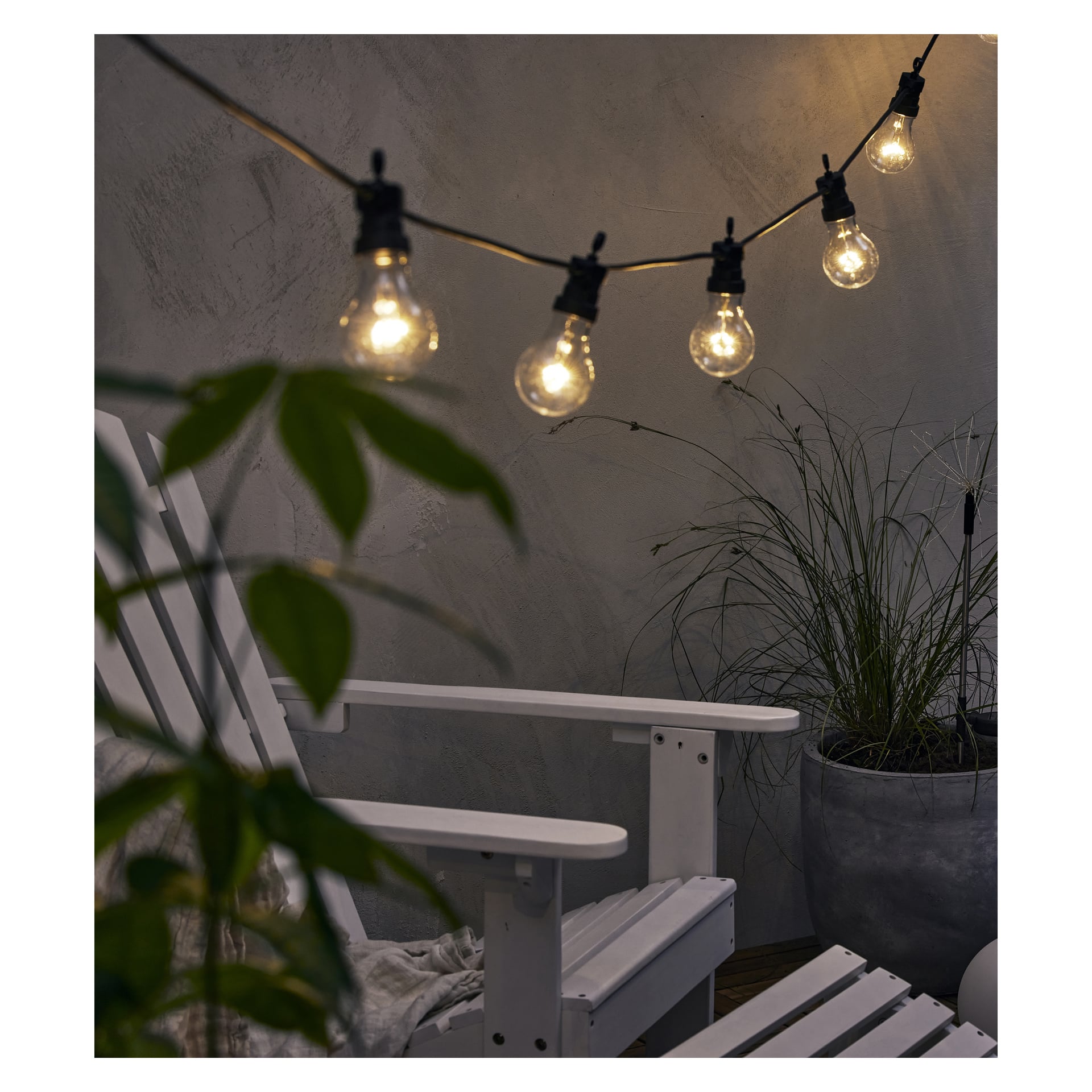 LED-Anslut-Partylichter mit runden Glühbirnen hängen über einem weißen Stuhl und Topfpflanzen an einer grau strukturierten Wand für ein gemütliches Gefühl.