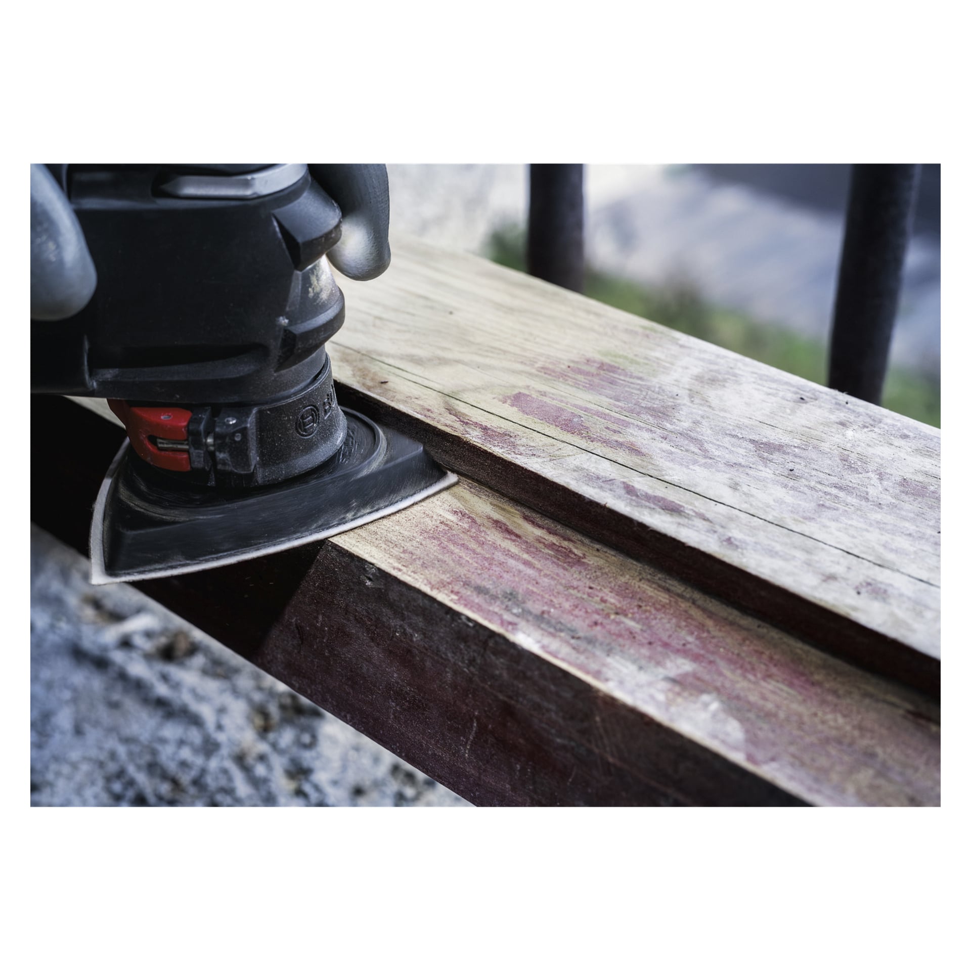 En person med handske slipar en träplanka utomhus på en stenavsats med Bosch Pro Delta slipplatta AVZ93G.