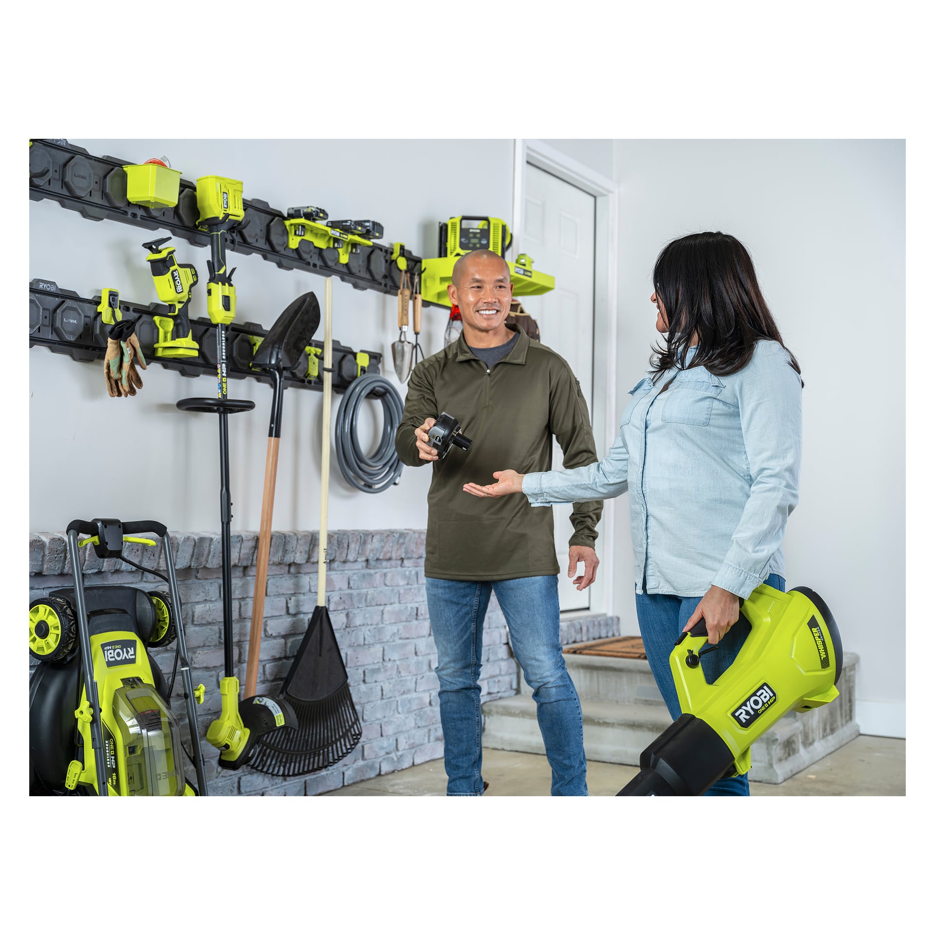 Två personer i ett garage med Ryobi LINK-räls; en håller ett batteri, den andra en fläkt, verktyg och utrustning prydligt på väggen.