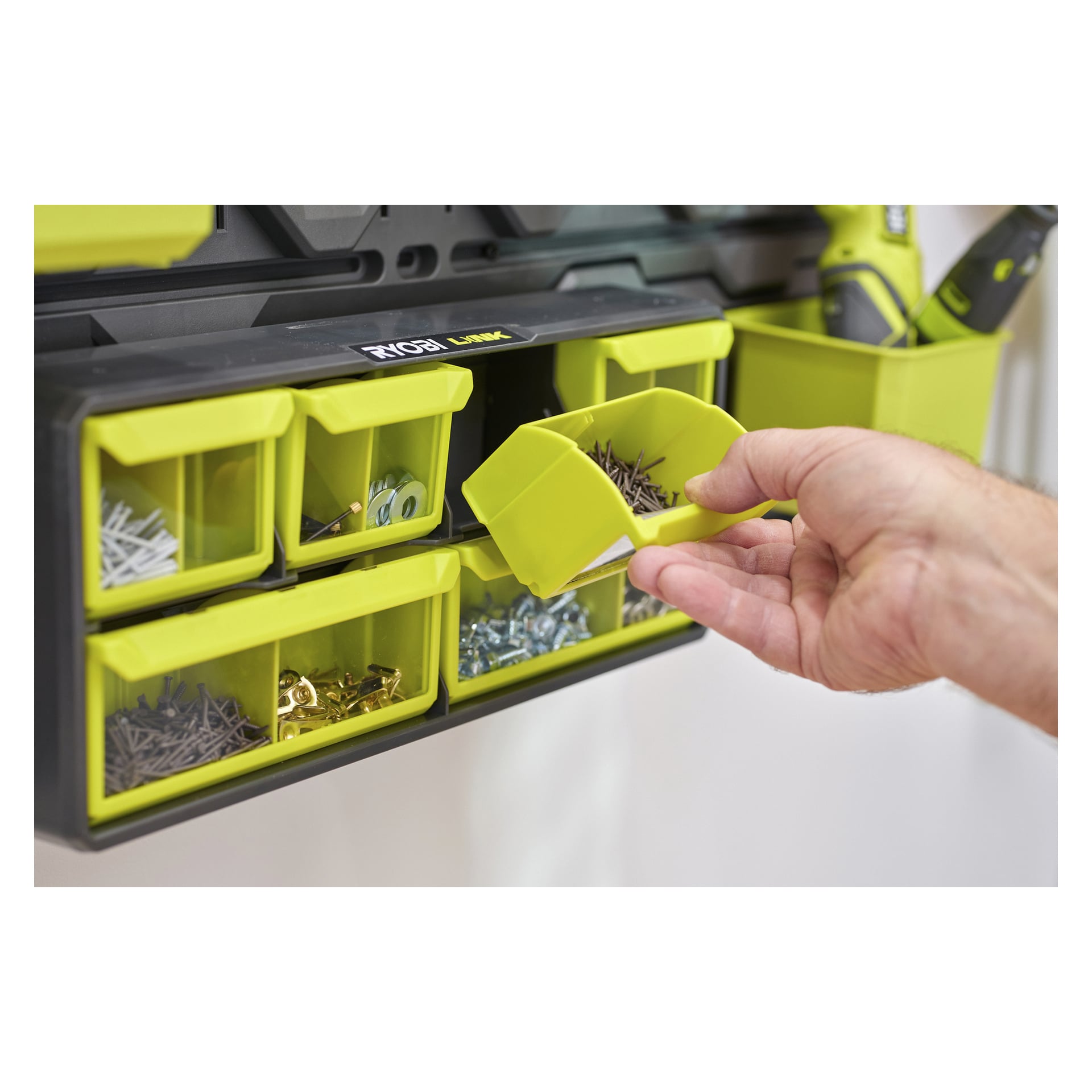 En hand tar bort en grön spiklåda från en Ryobi LINK-organiserare som håller fast låsen; Ett elverktyg är i bakgrunden.