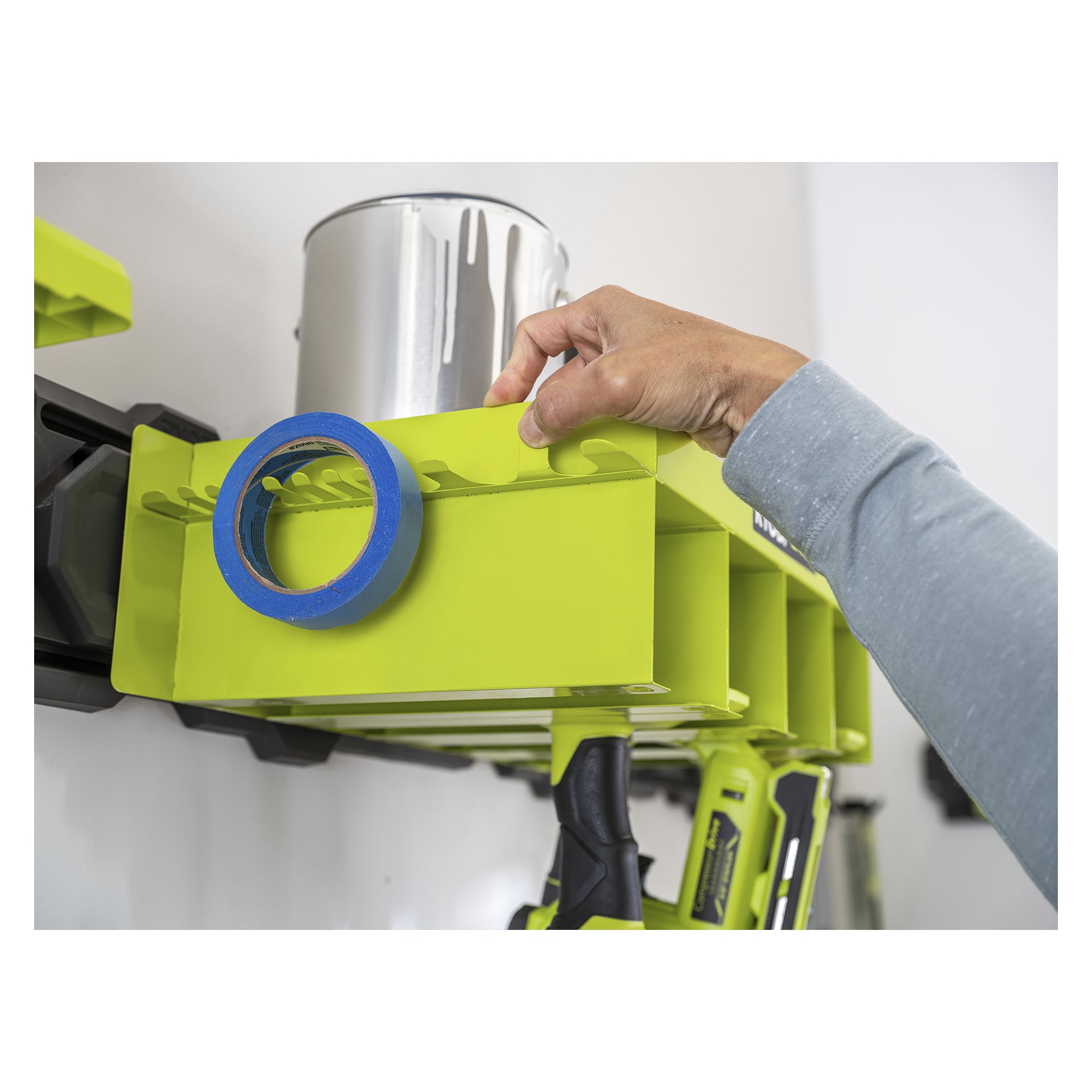 En person placerar en färgburk på en limegrön Ryobi LINK RSLW401 verktygshängare, med blå målartejp och sladdlösa verktyg under.