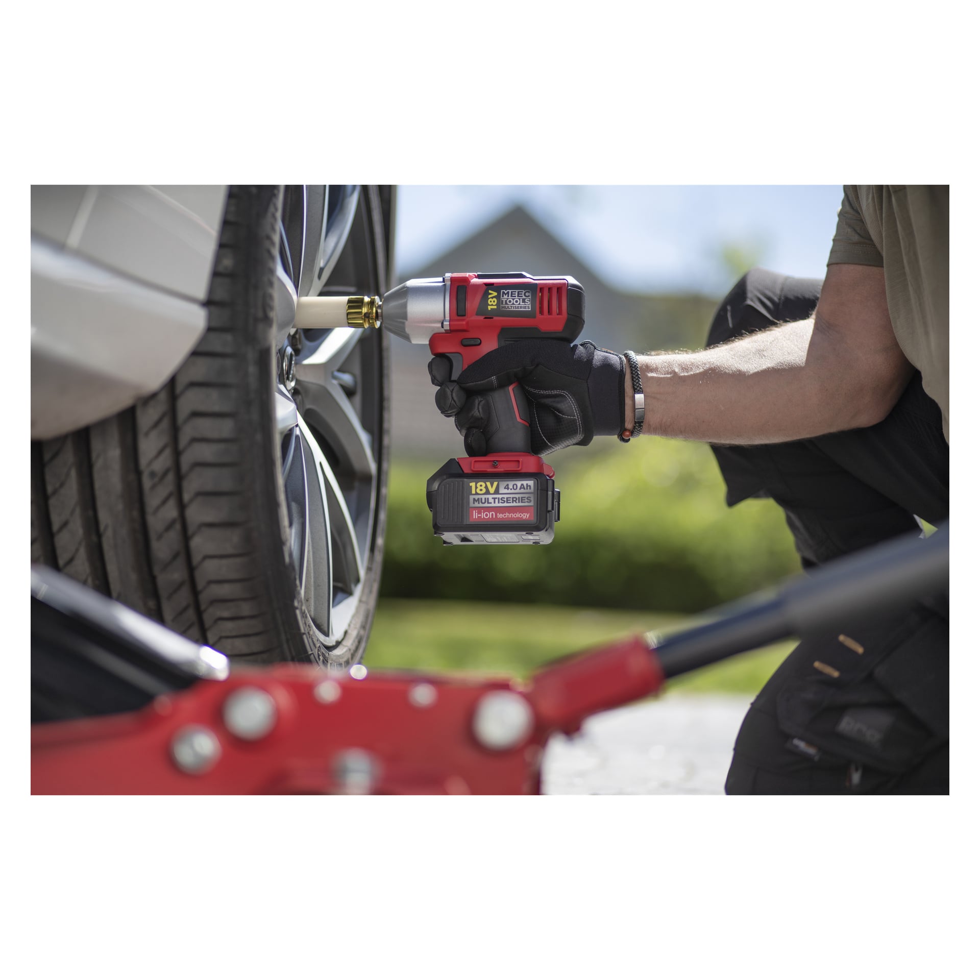 En person med handske använder en Meec Tools IW-18V EASY 100 slagnyckel på bilens hjulmutter, röd hydraulisk domkraft fram.