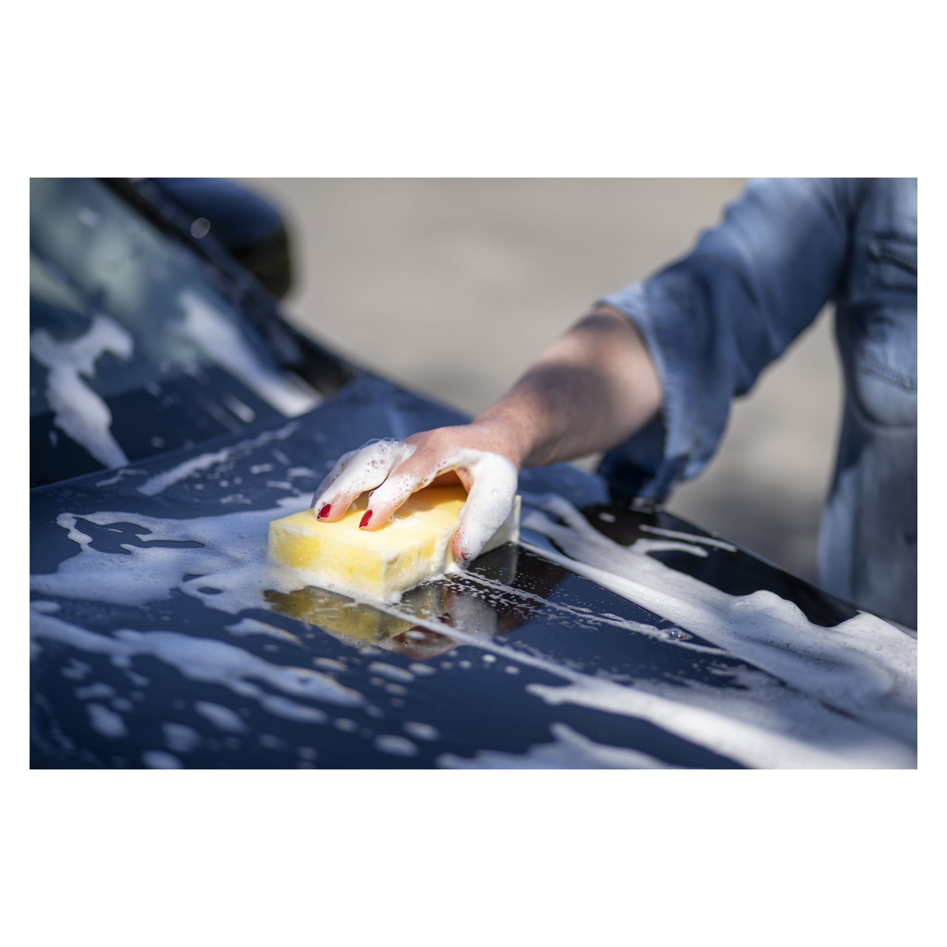 En person med rödmålade naglar, klädd i en jeansskjorta, rengör motorhuven på en svart bil med en Hamron Sponges med 10-pack schampo och sprider tvålskum.