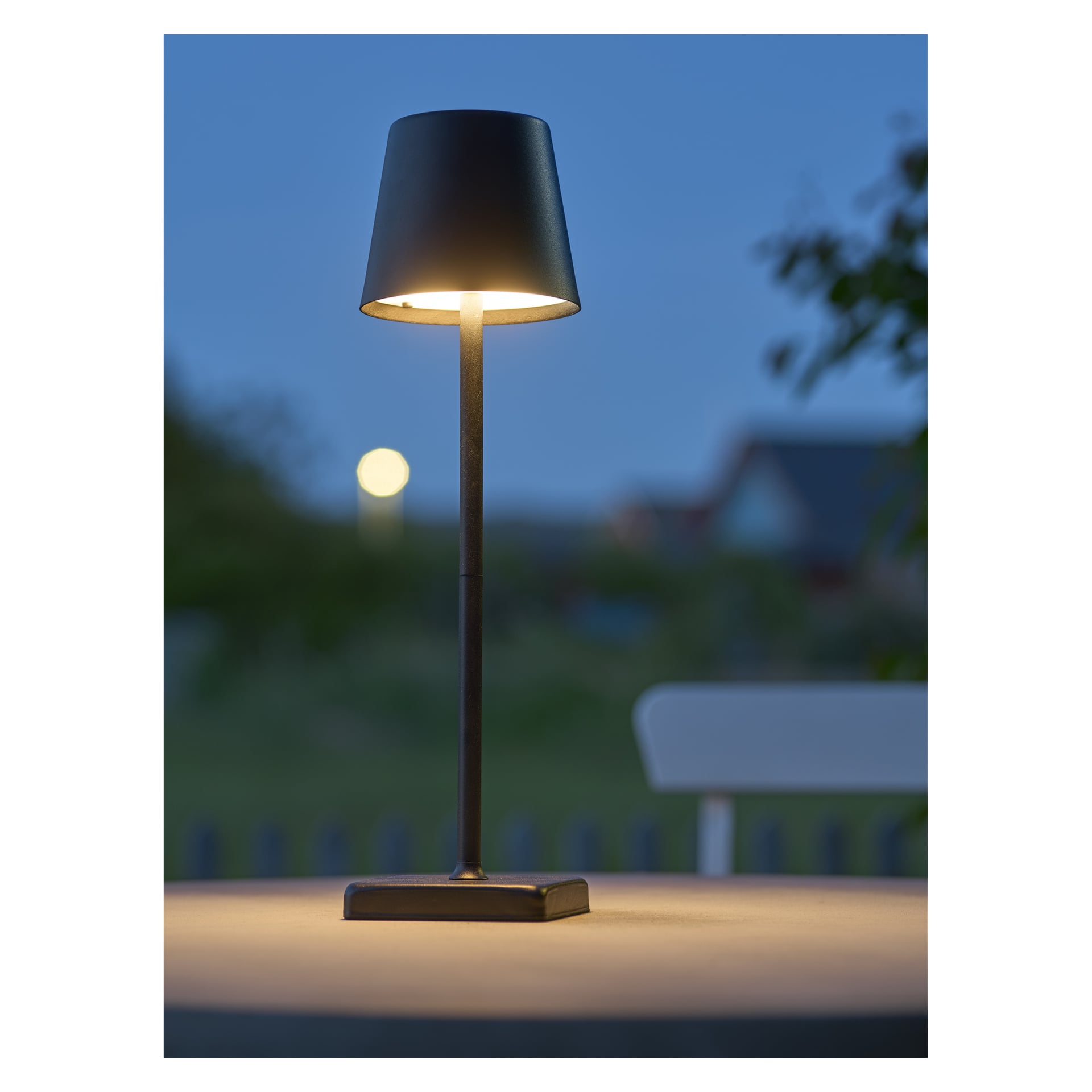 En solcellsdriven Anslut uppladdningsbar bordslampa lyser svagt i skymningen på ett utebord, svart konisk skärm lyser upp, grönska bakom.