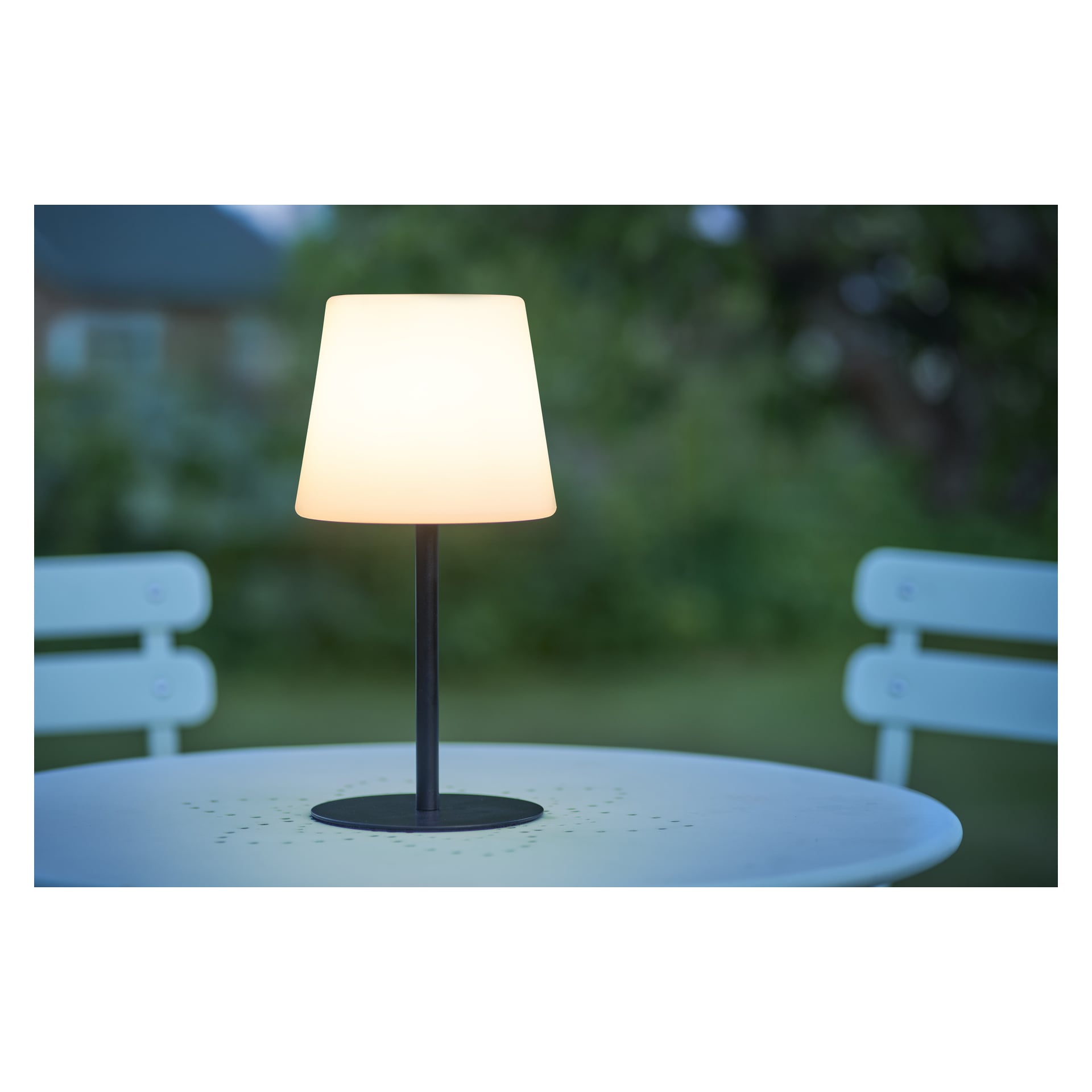 En uppladdningsbar bordslampa med solcells-LED från Anslut är placerad, tänd, på ett runt utebord mellan två vita stolar, med en bakgrund av suddig grönska.