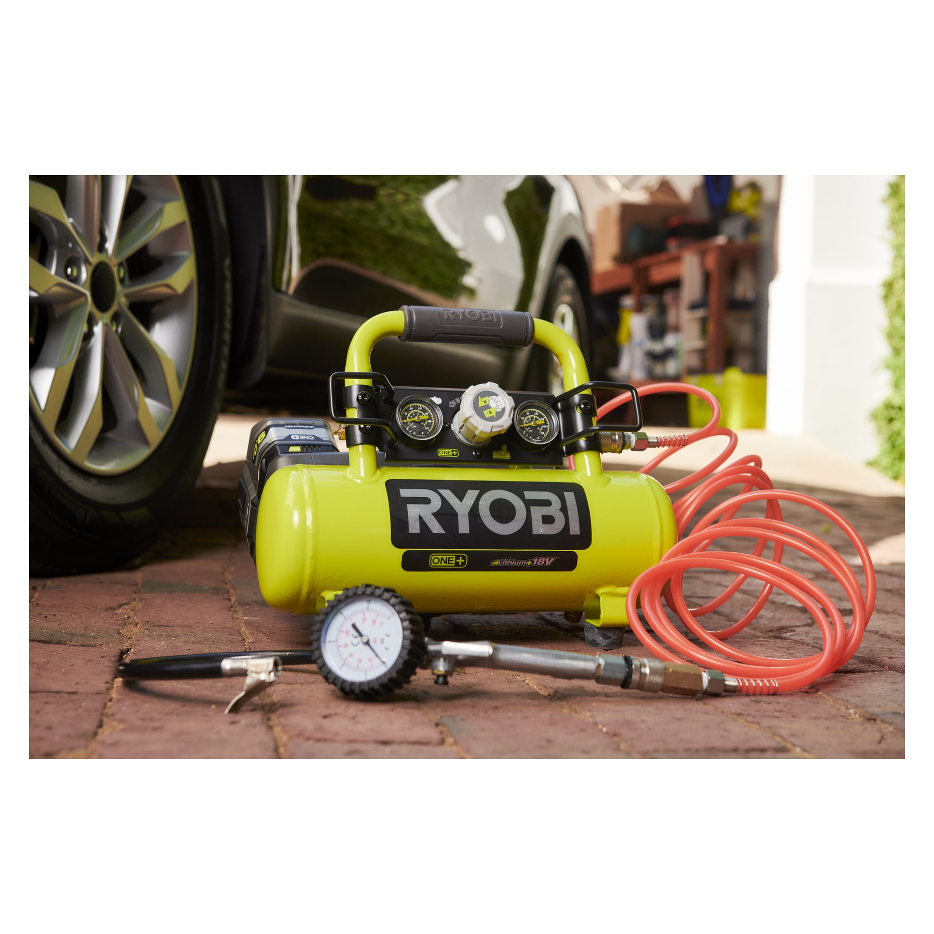 Gul Ryobi One+ R18AC-0 kompressor med handtag och orange slang står på tegelstenar nära en bil; Verktyg och en tryckmätare ligger i närheten.