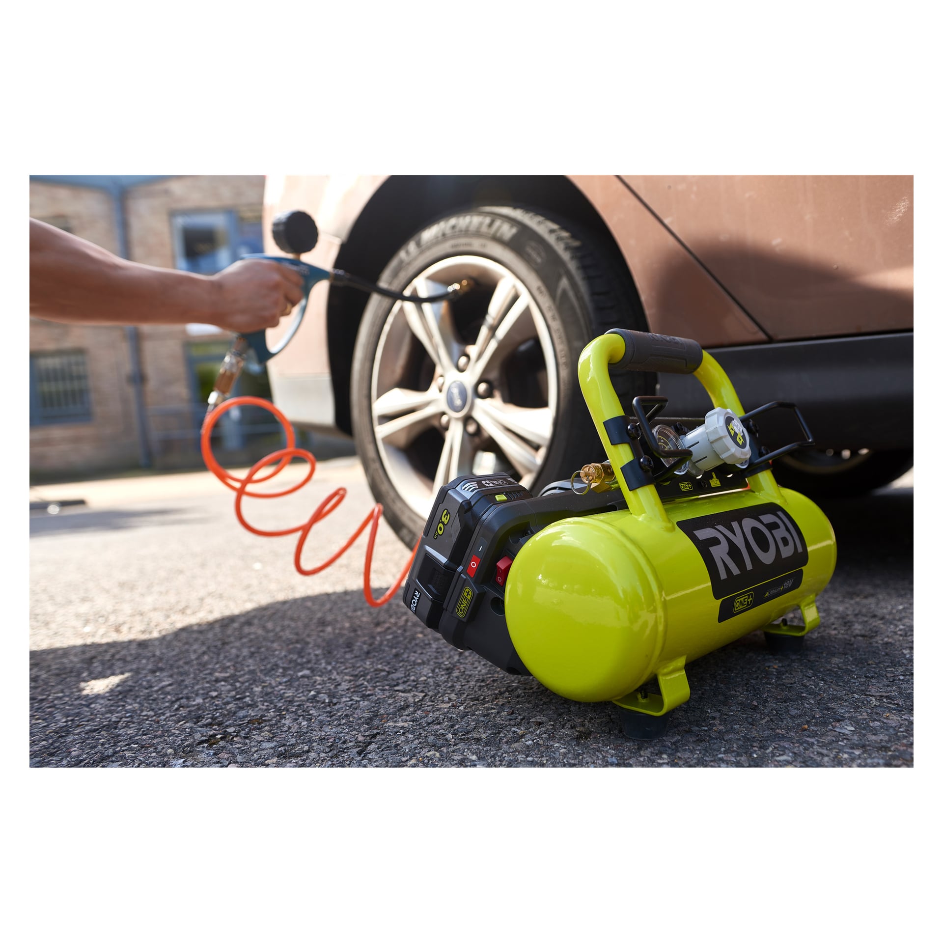 En person fyller upp ett brunt bildäck med en Ryobi One+ R18AC-0 batterikompressor och orange slang på en asfalterad yta.