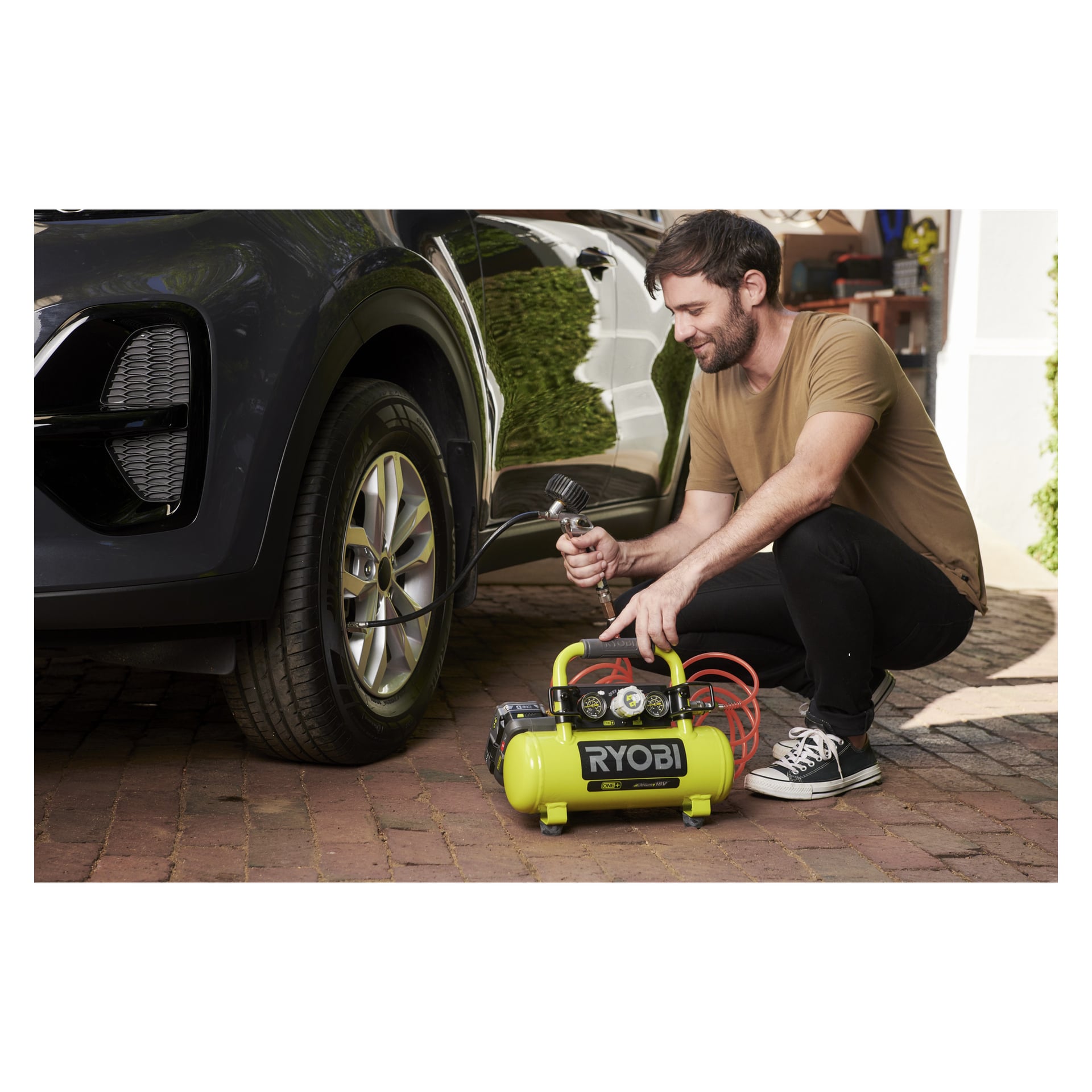 En man hukar på en tegeluppfart och pumpar upp ett bildäck med en Ryobi One+-kompressor; Verktyg är utspridda i bakgrunden.