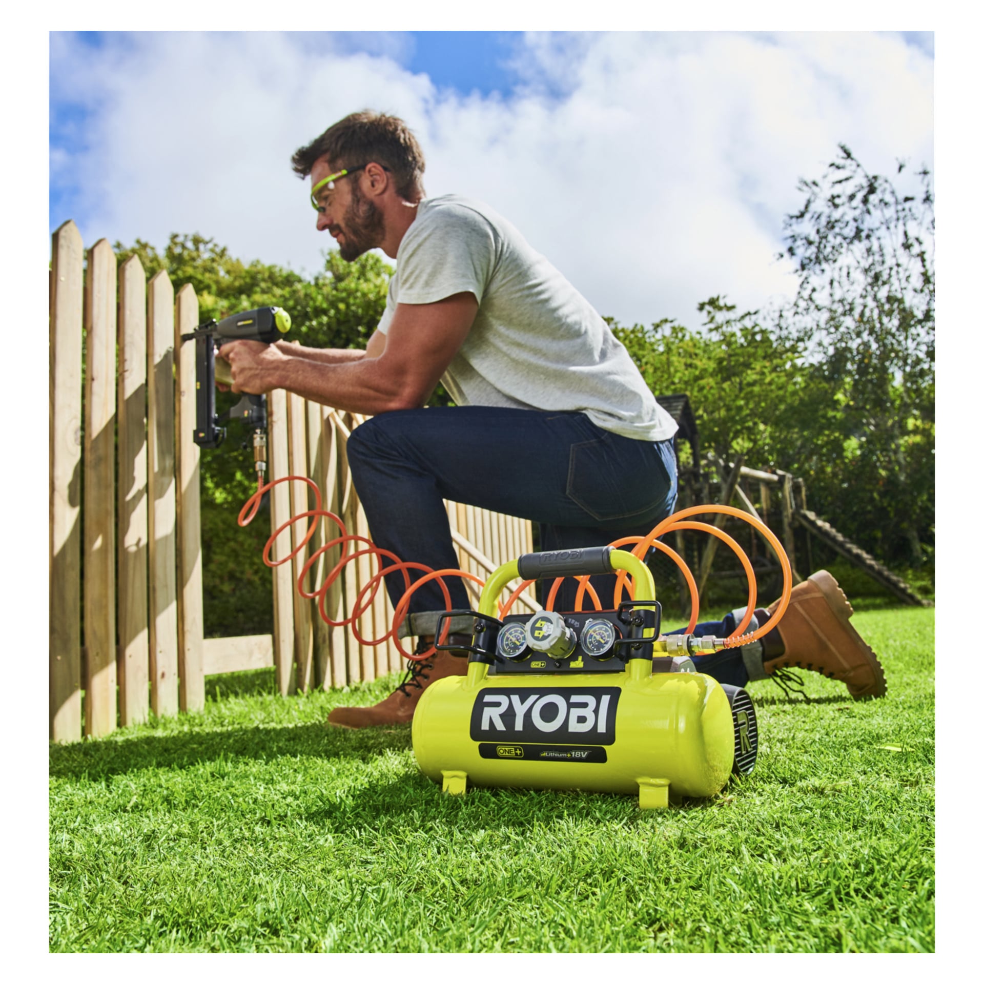En man med skyddsglasögon knäböjer på gräset och använder en Ryobi One+ kompressor och spikpistol för att fästa trästängsel med en orange slang.