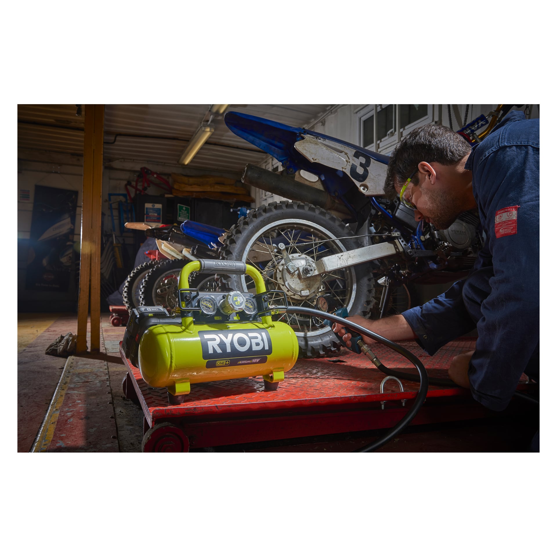 Någon knäböjer vid en crossmotorcykel på en garageplattform och använder en Ryobi One+ R18AC-0-kompressor för att pumpa däcket; Verktyg och en annan cykel dyker upp bakom.