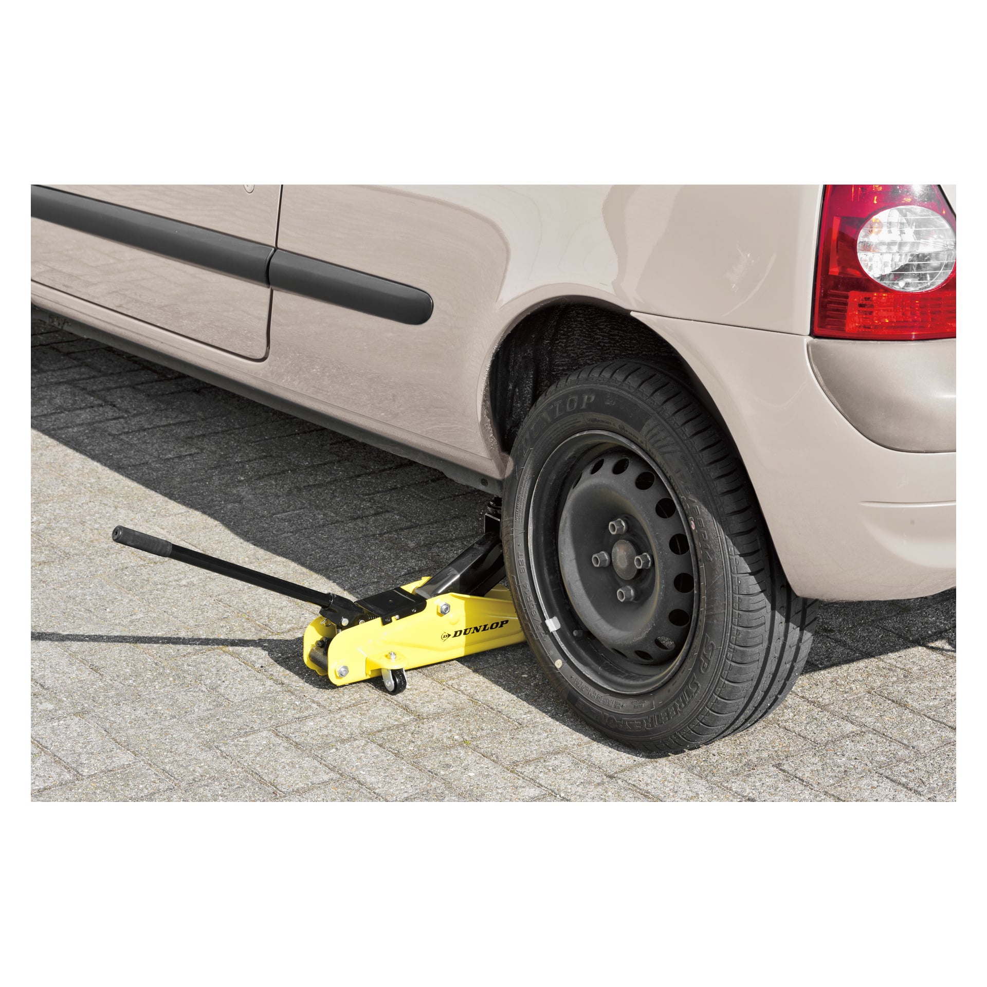 En gul Dunlop hydraulisk jekk løfter bakenden på en beige bil og løfter det ene hjulet opp fra bakken på et asfaltert underlag.