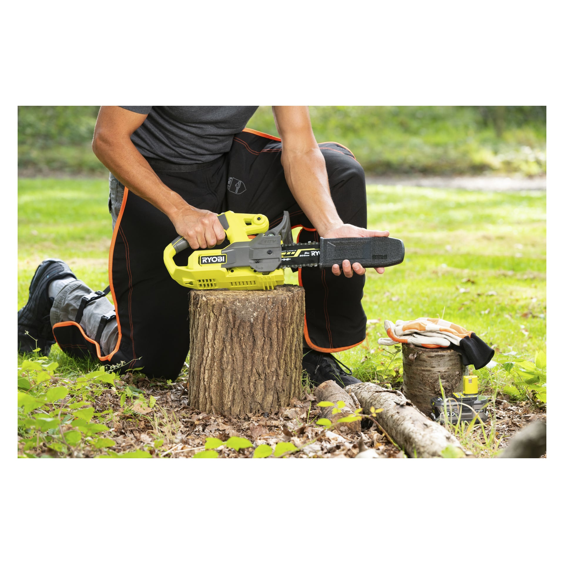 Noen kneler på gresset og fester et svart bladdeksel til en gul Ryobi One+ motorsag på en stubbe, hansker og batteri ved siden av.
