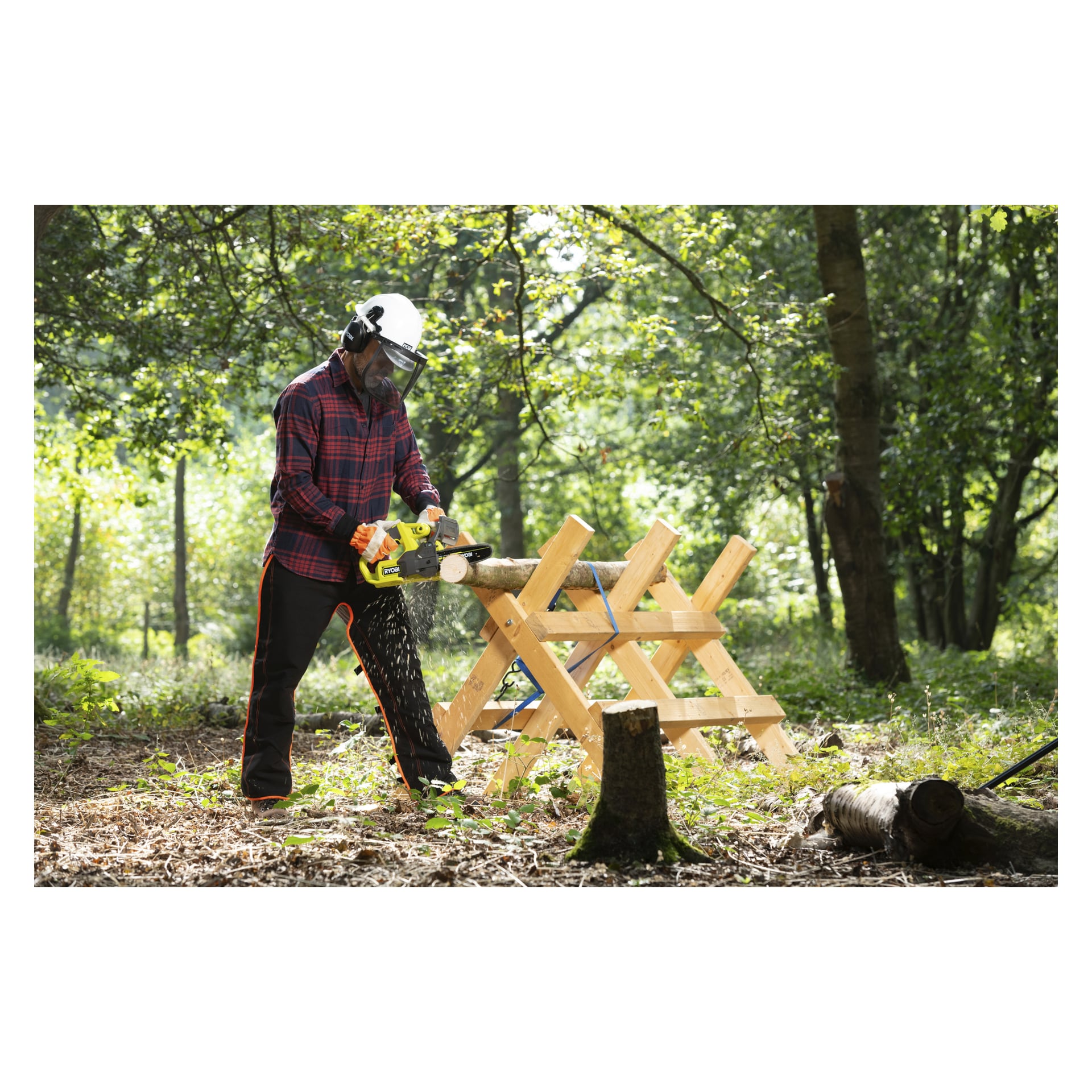 En person i sikkerhetsutstyr kutter en stokk på en sagbukk med en Ryobi One+ batterimotorsag i solfylt skog mens flisene flyr.