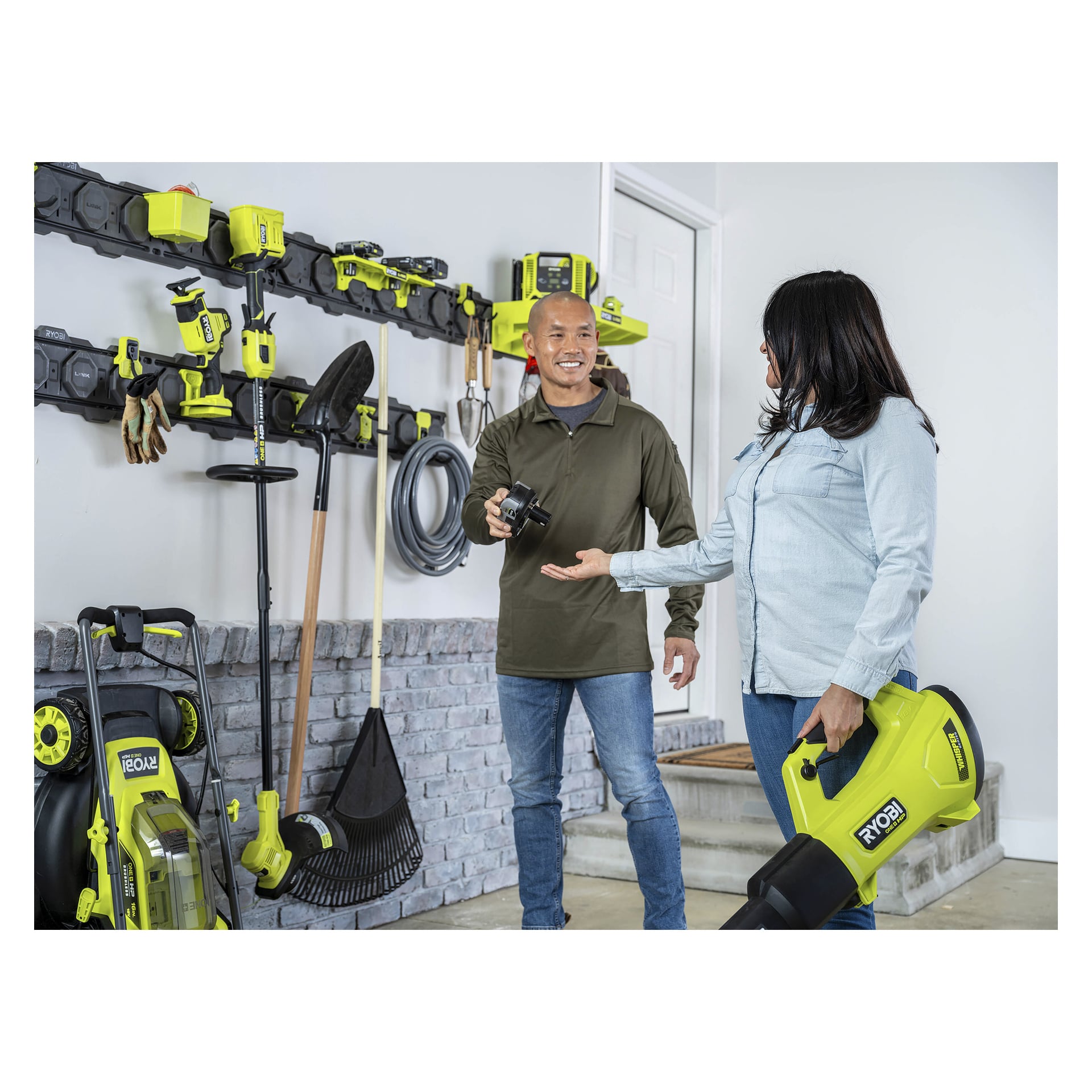 En man och en kvinna står i ett prydligt garage med Ryobi Starter-kit LÄNK RSLWPK-8PCJ 8 delar. Mannen håller i ett batteri och kvinnan har en lövblåsare. Bakom dem är verktyg prydligt uppställda på väggställningar.
