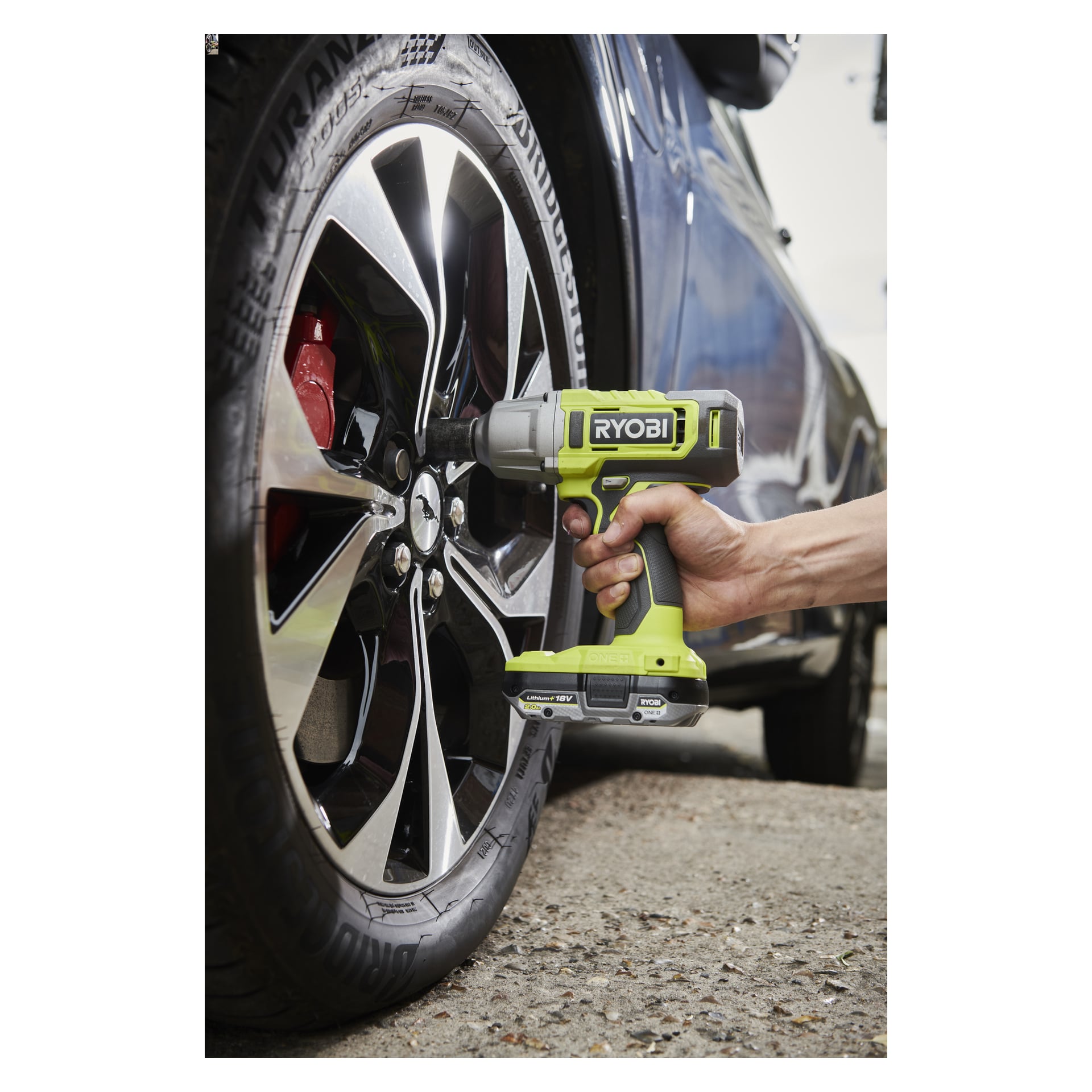 Noen bruker en Ryobi One+ Impact Wrench Kit RIW18-1C20SA3 på hjulmutterne til en mørk bil utstyrt med Bridgestone Turanza-dekk.