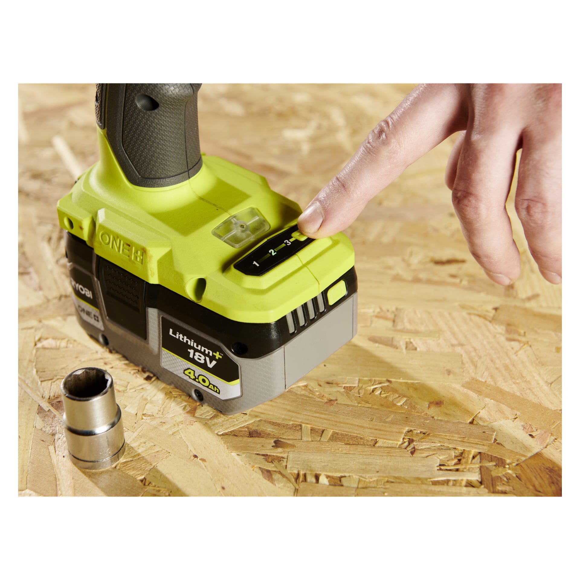 En hånd sjekker batterinivået på en Ryobi One+ Impact Wrenchs 18V-batteri, festet til verktøyet, liggende på tre ved en kontakt.