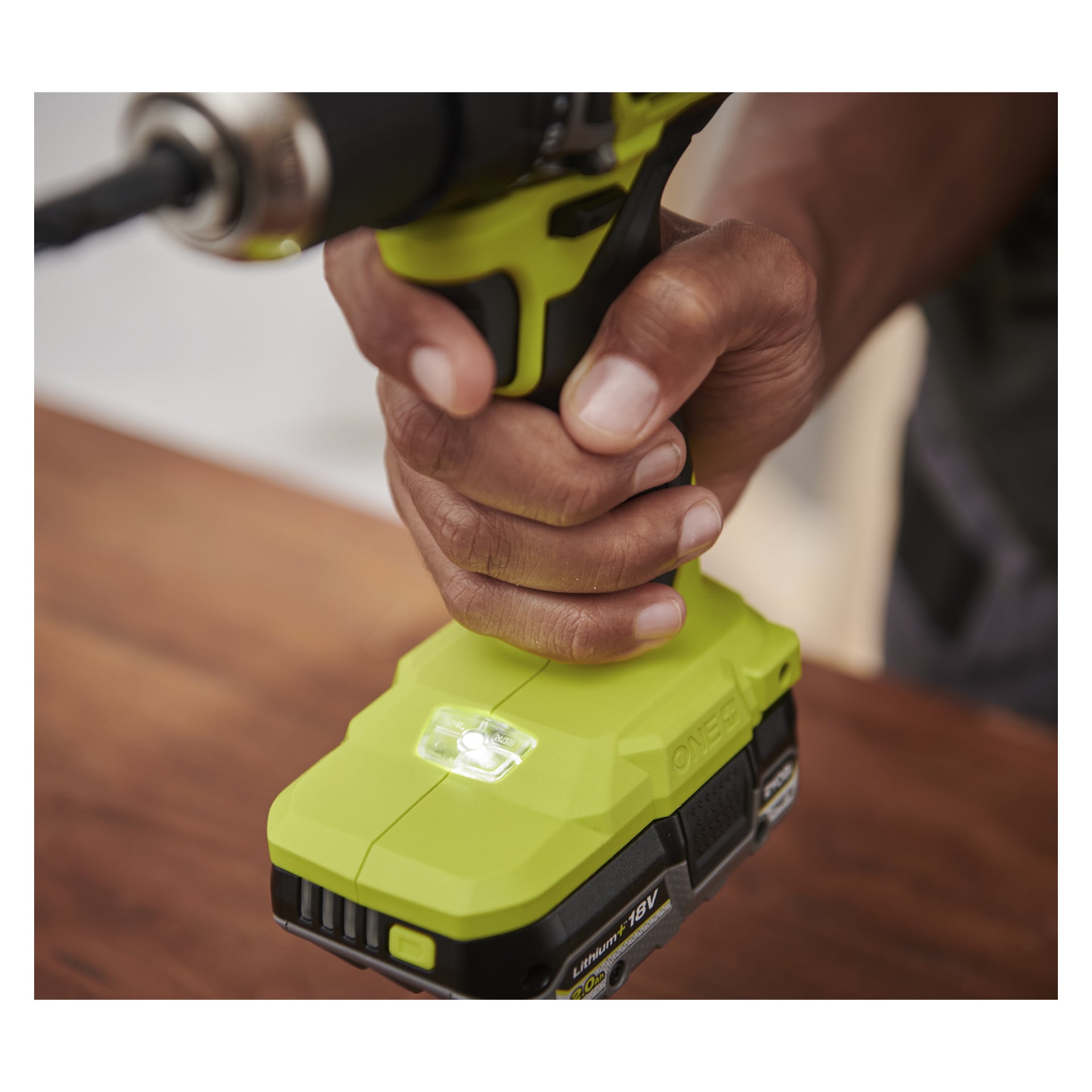 Någon håller i ett Ryobi One+ HP borstlöst skruvmejselkit, ett grönt verktyg med svart-gult batteri och LED-lampa på träytan.