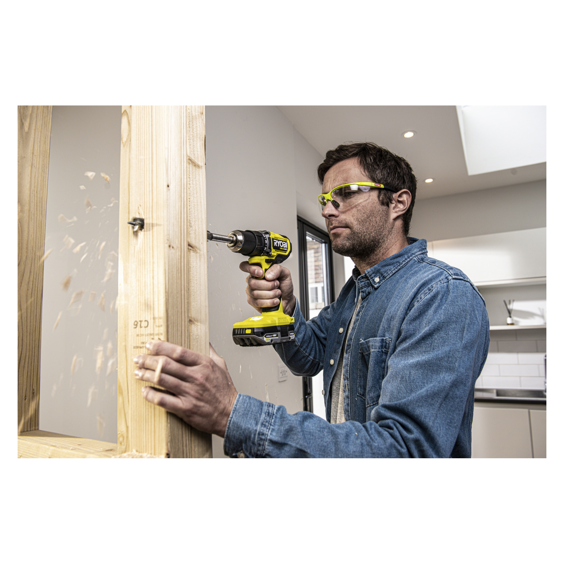 En person med skyddsglasögon och jeansskjorta borrar i en träram med en Ryobi One+ HP borstlös skruvmejsel inomhus.