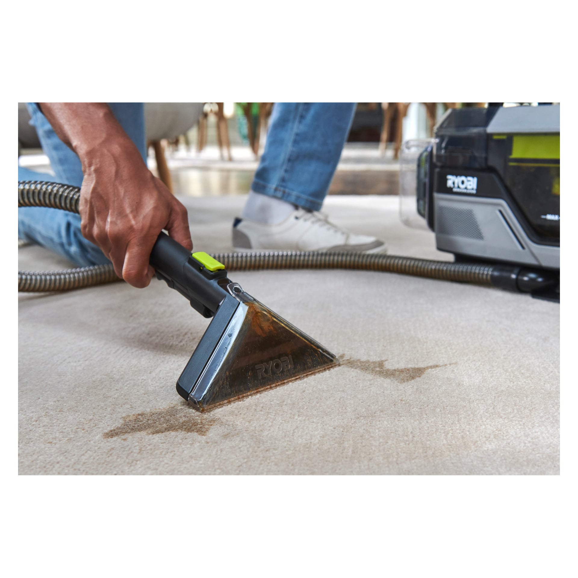 En person renser en flekk på et lett teppe med en Ryobi One+ Swift Clean batteridrevet flekkfjerner med gjennomsiktig dyse og slange.