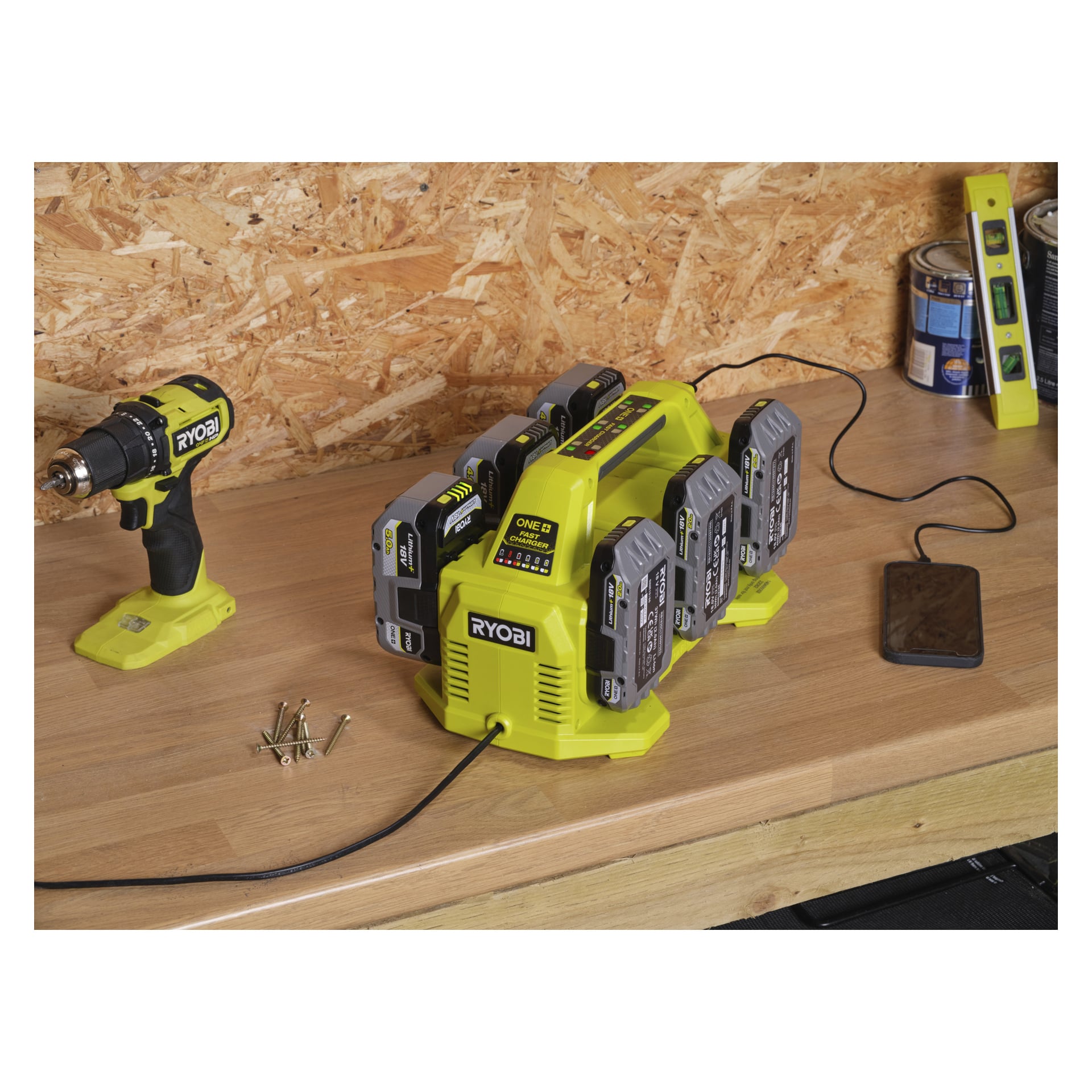 En Ryobi One+ Multilader med batterier står og lades på en arbeidsbenk i tre sammen med en drill, skruer, malingsbokser, telefon og vaterpas.