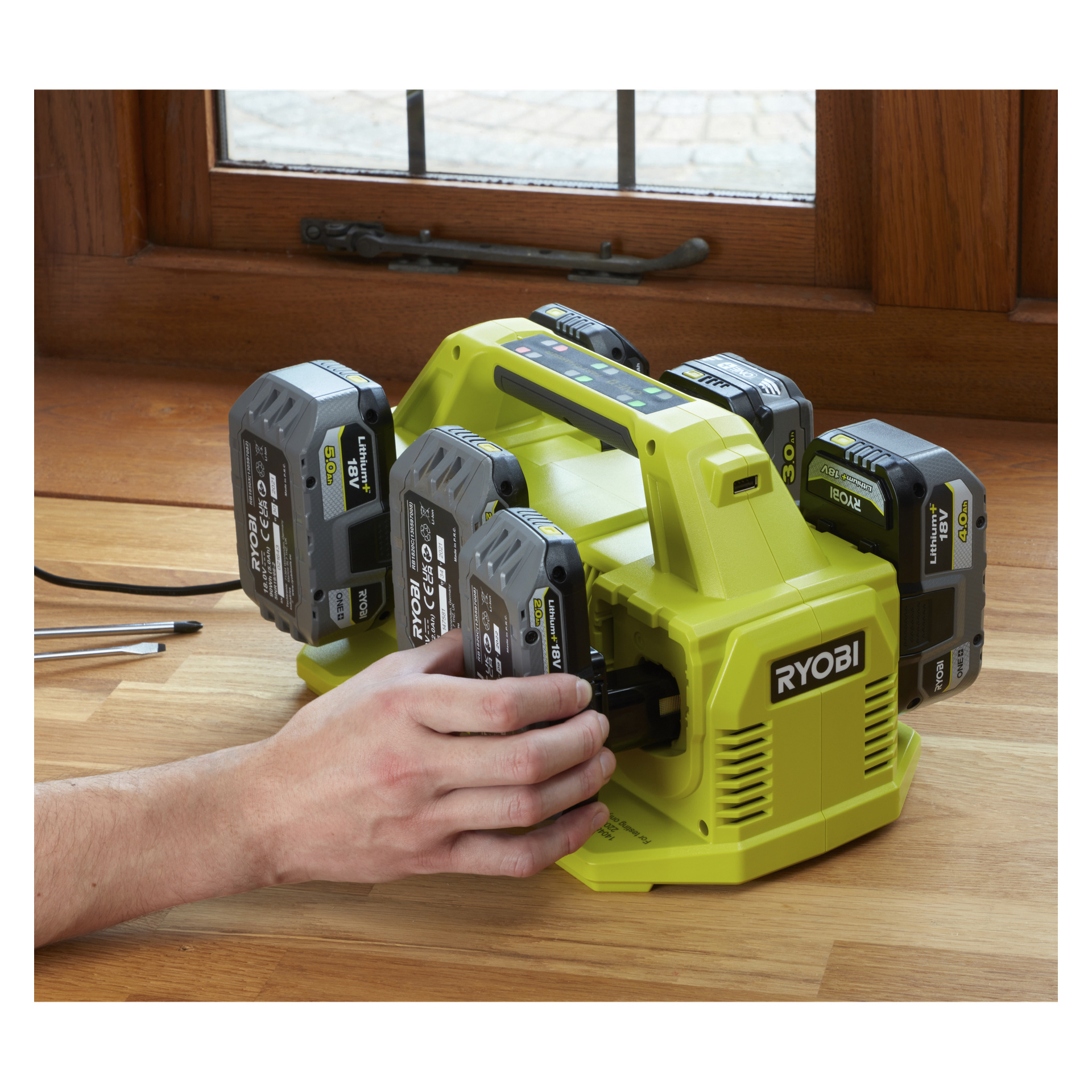 Noen plasserer en Ryobi One+ Multi-lader RC18640-3422 med fire batterier i en grønn ladestasjon ved et vindu på tre, med flere Ryobi-batterier festet.