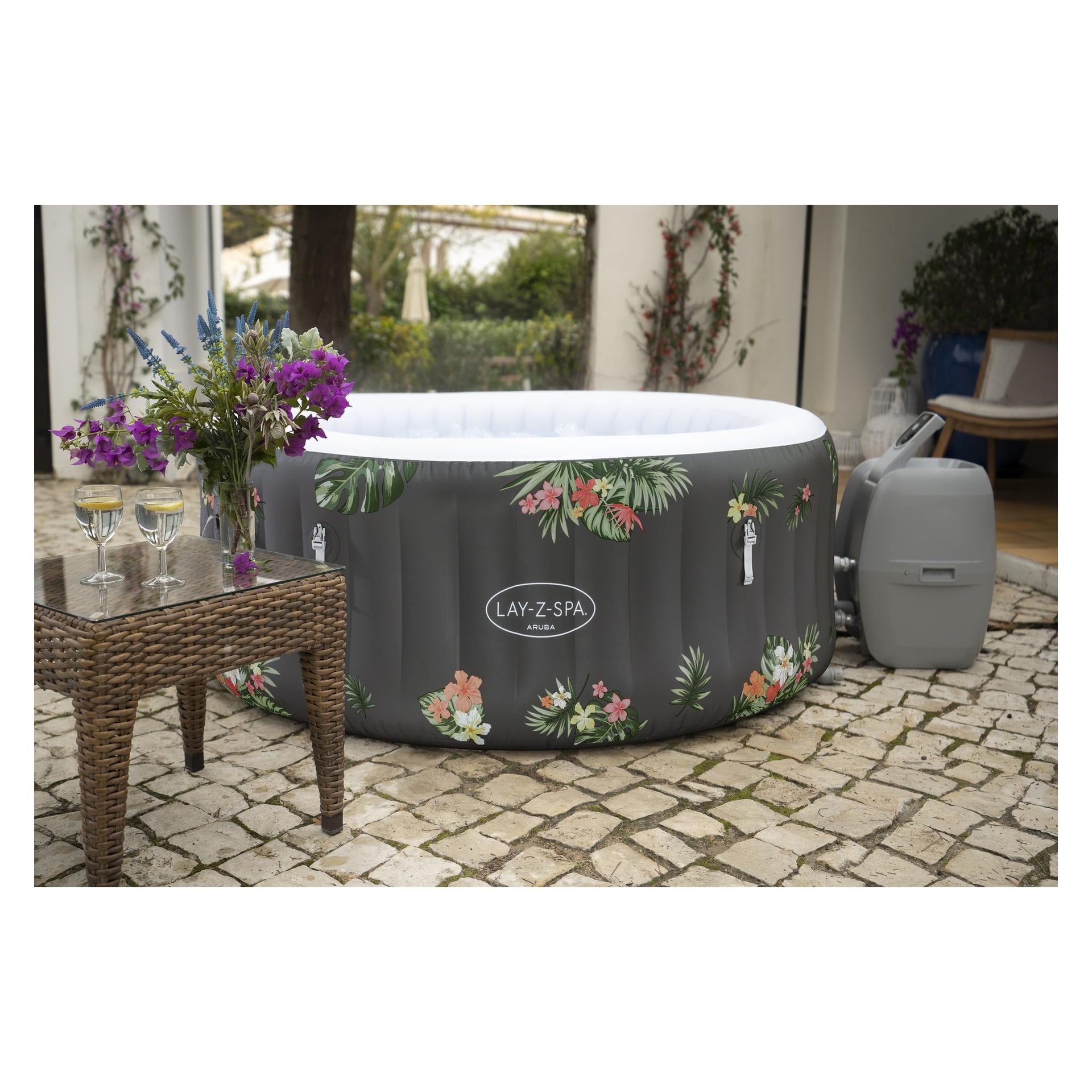 Tropikalny AirJet LAY-Z-SPA Aruba stoi na kamiennym patio obok wiklinowego stołu z fioletowymi kwiatami i kieliszkami do białego wina.