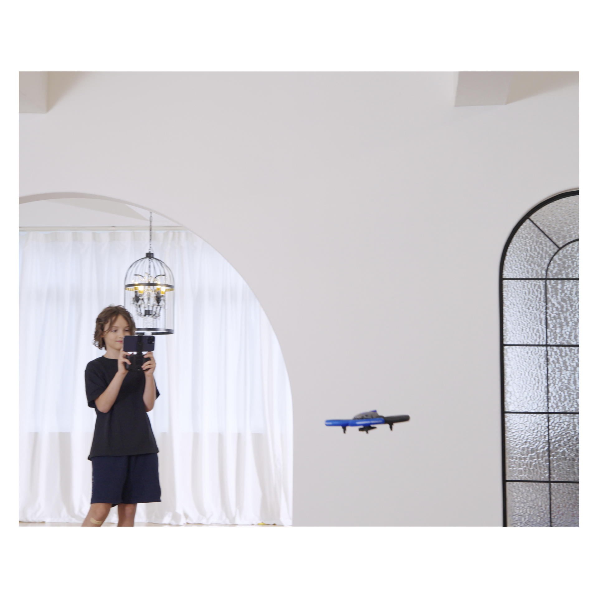 Dziecko w pomieszczeniu używa tabletu, aby pilotować mały niebieski Silverlit Bumper Drone Vision WiFi w pomieszczeniu z białymi ścianami i łukowatym oknem.